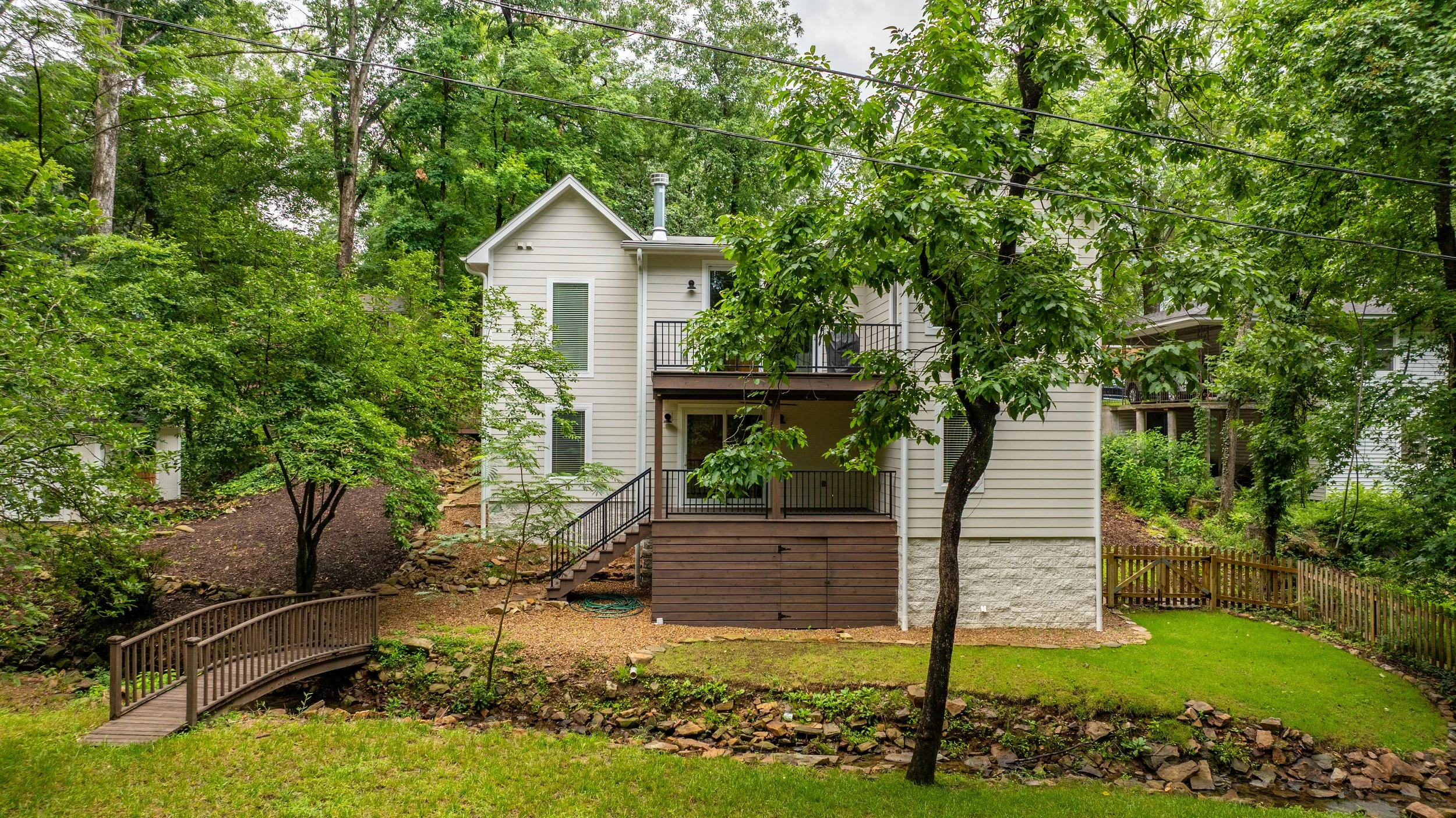 A two-story house with beige siding and a wooden deck on the second floor, surrounded by lush green trees and a well-maintained yard with a small wooden bridge crossing a dry creek bed.
