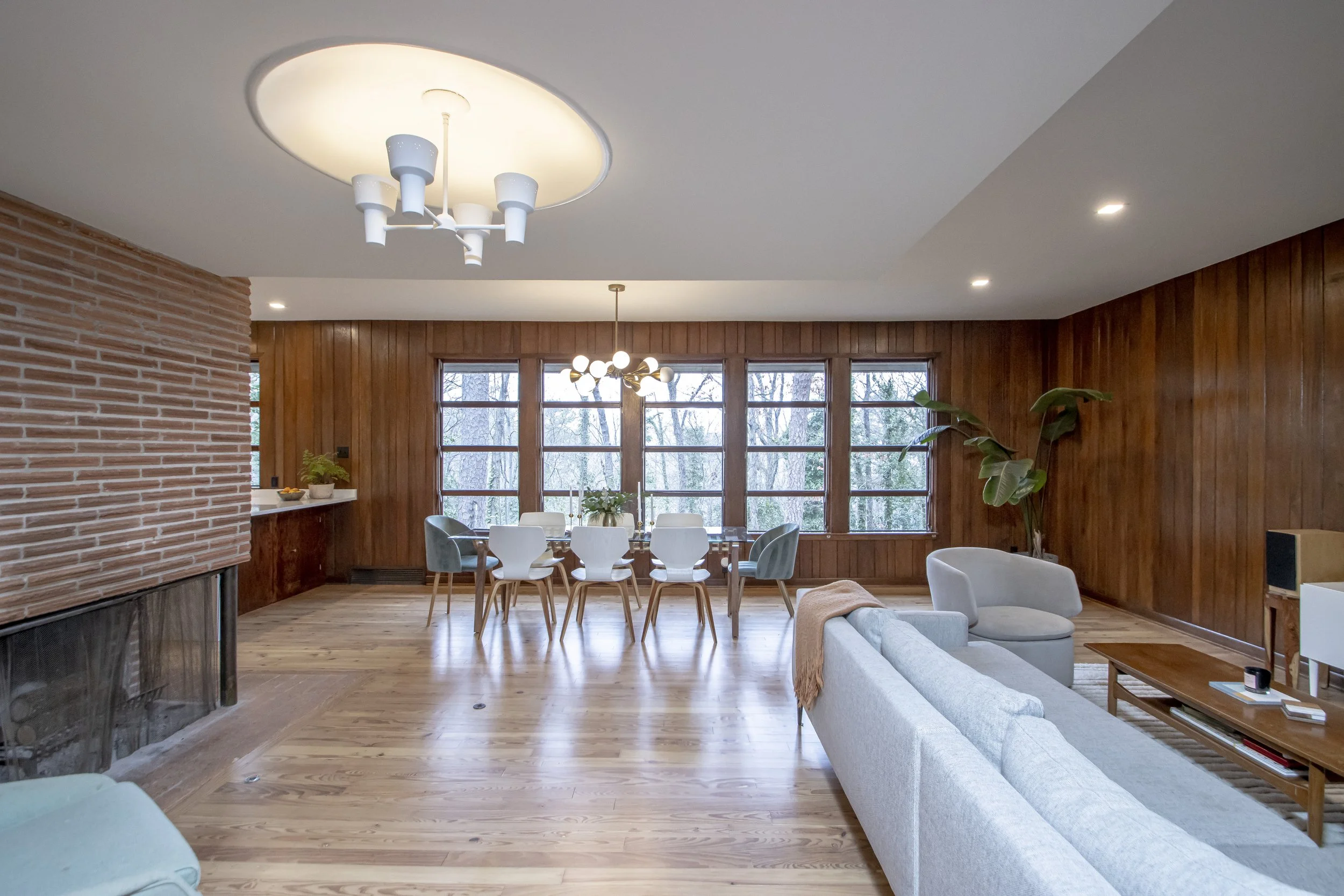 A Mid-century Modern style living room with wood tall panel walls, three large picture windows overlooking the woods, and a white ceiling with two modern light fixtures. There is a light gray upholstered sofa and a matching armchair, a wooden coffee 