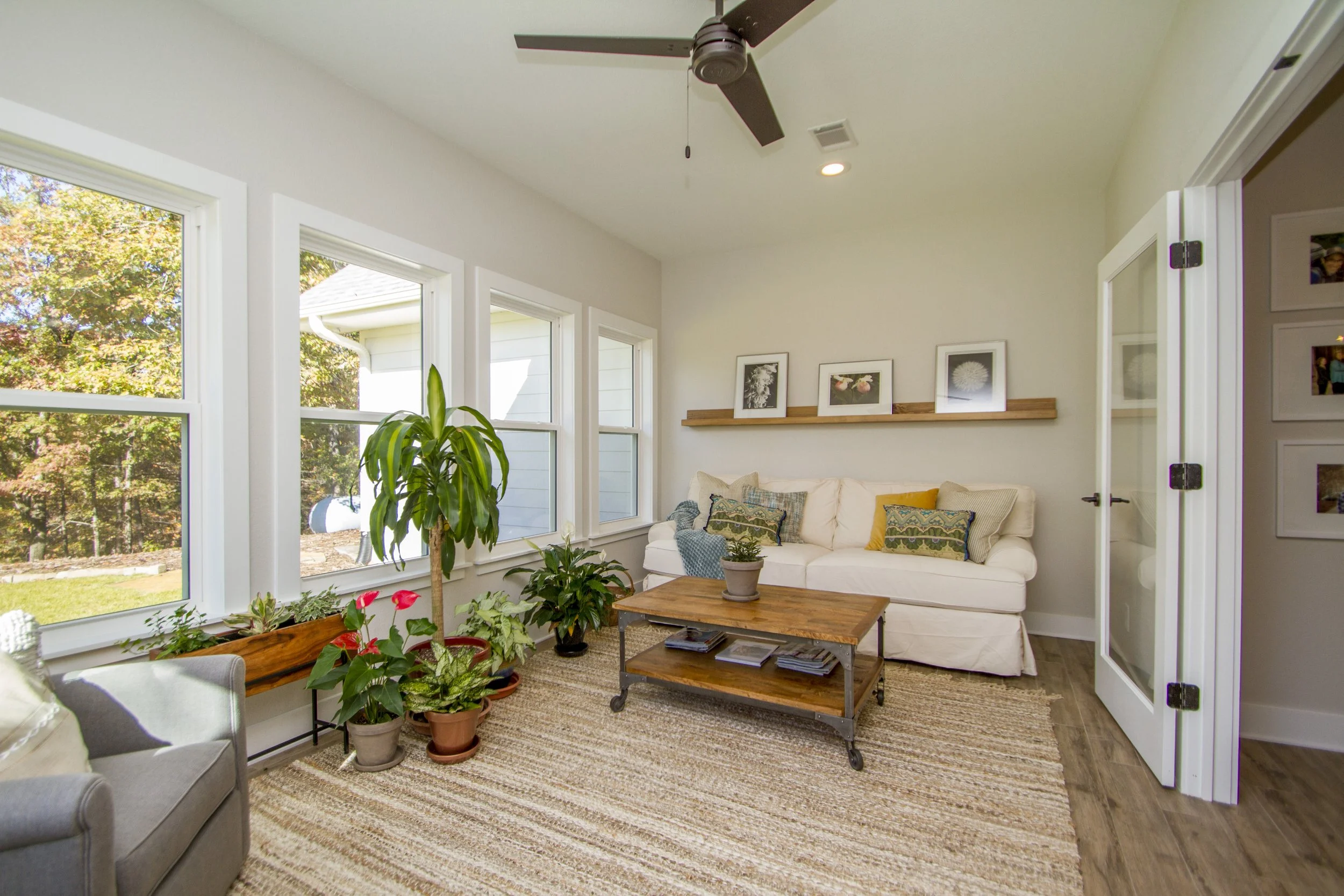 Living room with beige sofa, wooden coffee table, potted plants, large windows showing trees and house, and a ceiling fan.