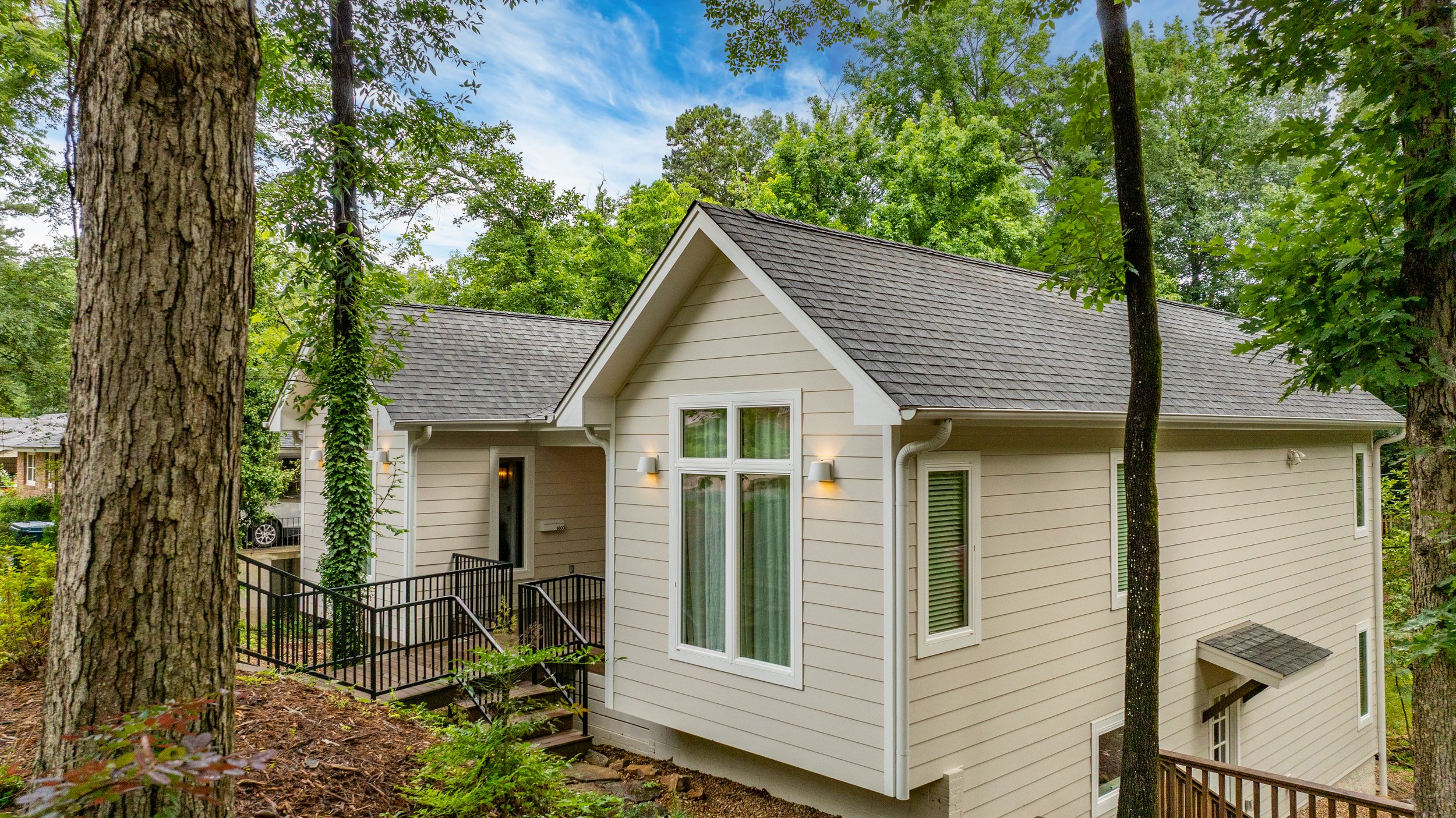A beige house surrounded by tall trees in a wooded area, with large windows, a small outdoor staircase, and exterior lights on.