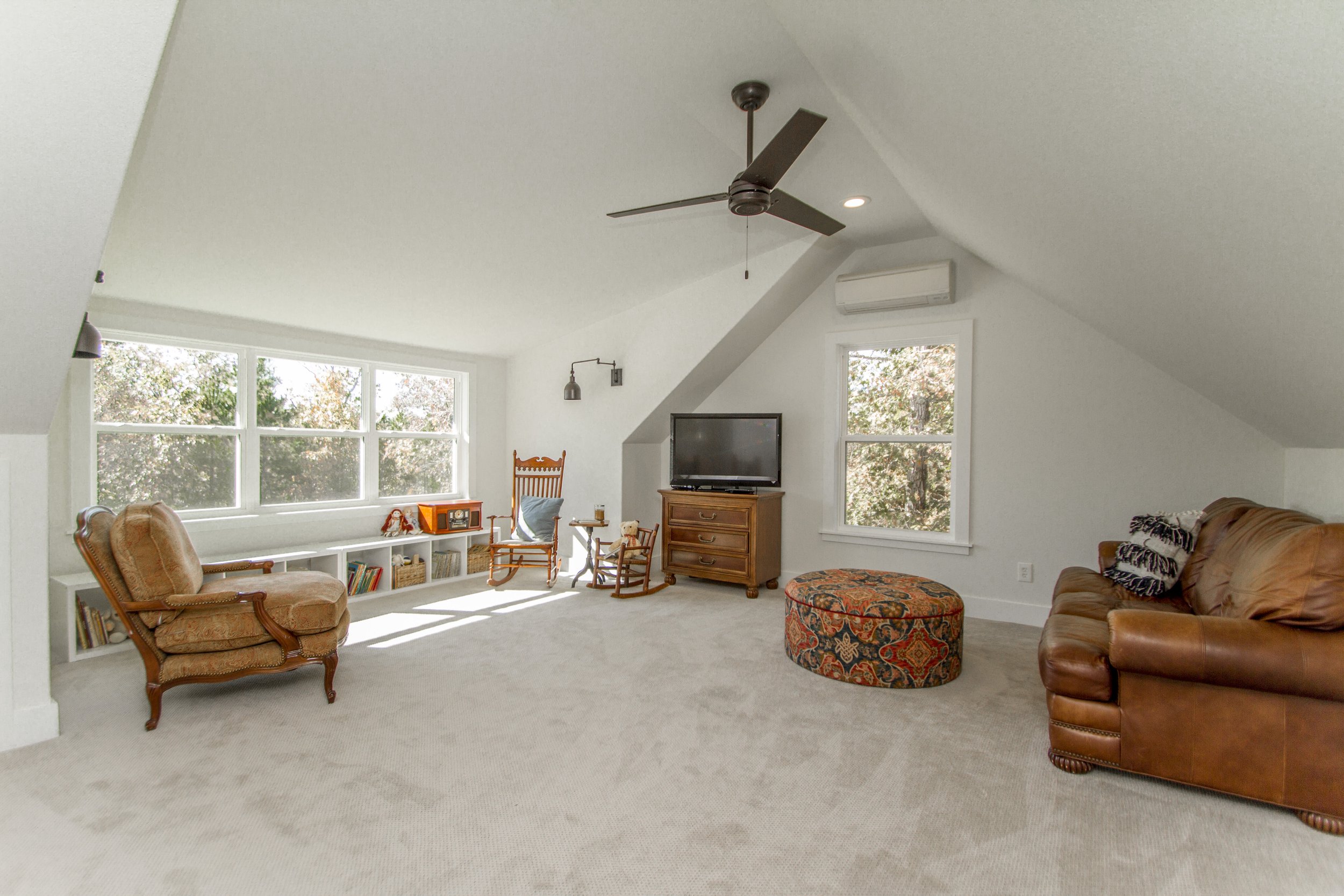 Living room with large windows, beige carpet, leather and wooden furniture, a TV on a wooden stand, and a ceiling fan.