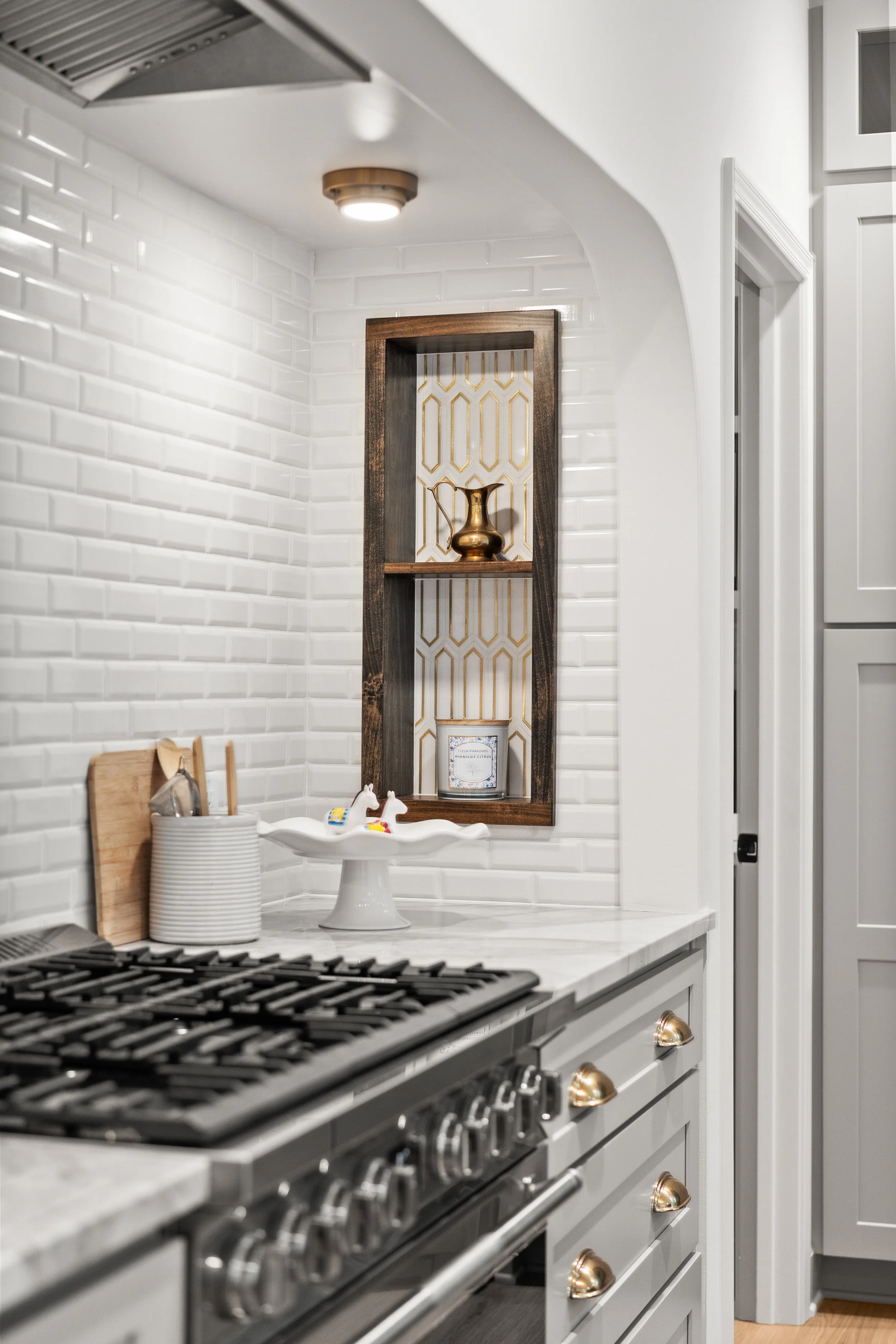 A modern kitchen featuring white subway tile backsplash, a gorilla glass stove with black grates, white cabinetry with gold knobs, and a decorative wooden shelf with a gold vase, framed photo, and patterned backsplash behind the shelf.