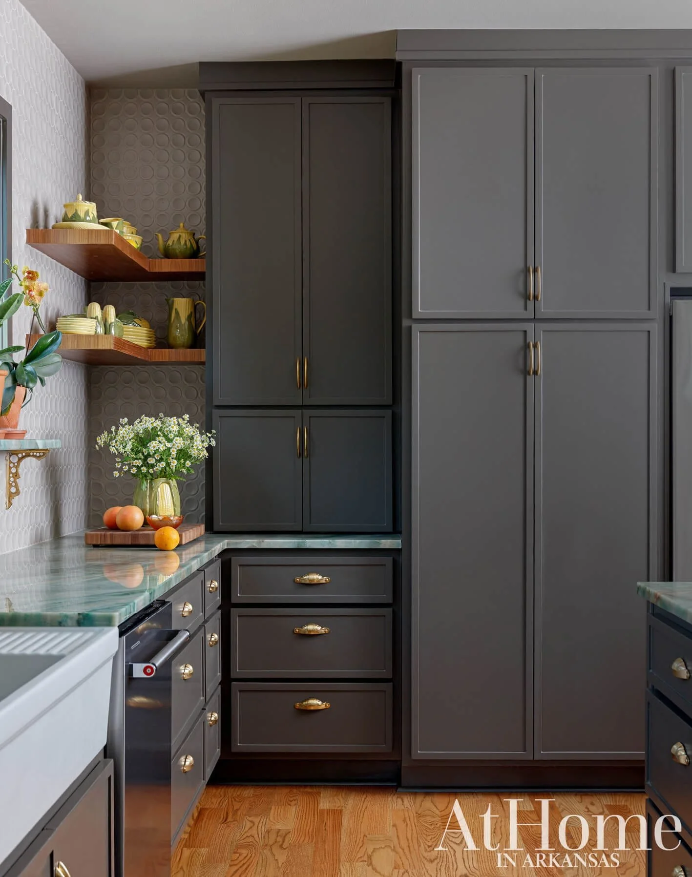 Kitchen with dark gray cabinets, open wooden shelves with green tea set, marble countertop with oranges and white flowers, patterned white wall, and hardwood floor.