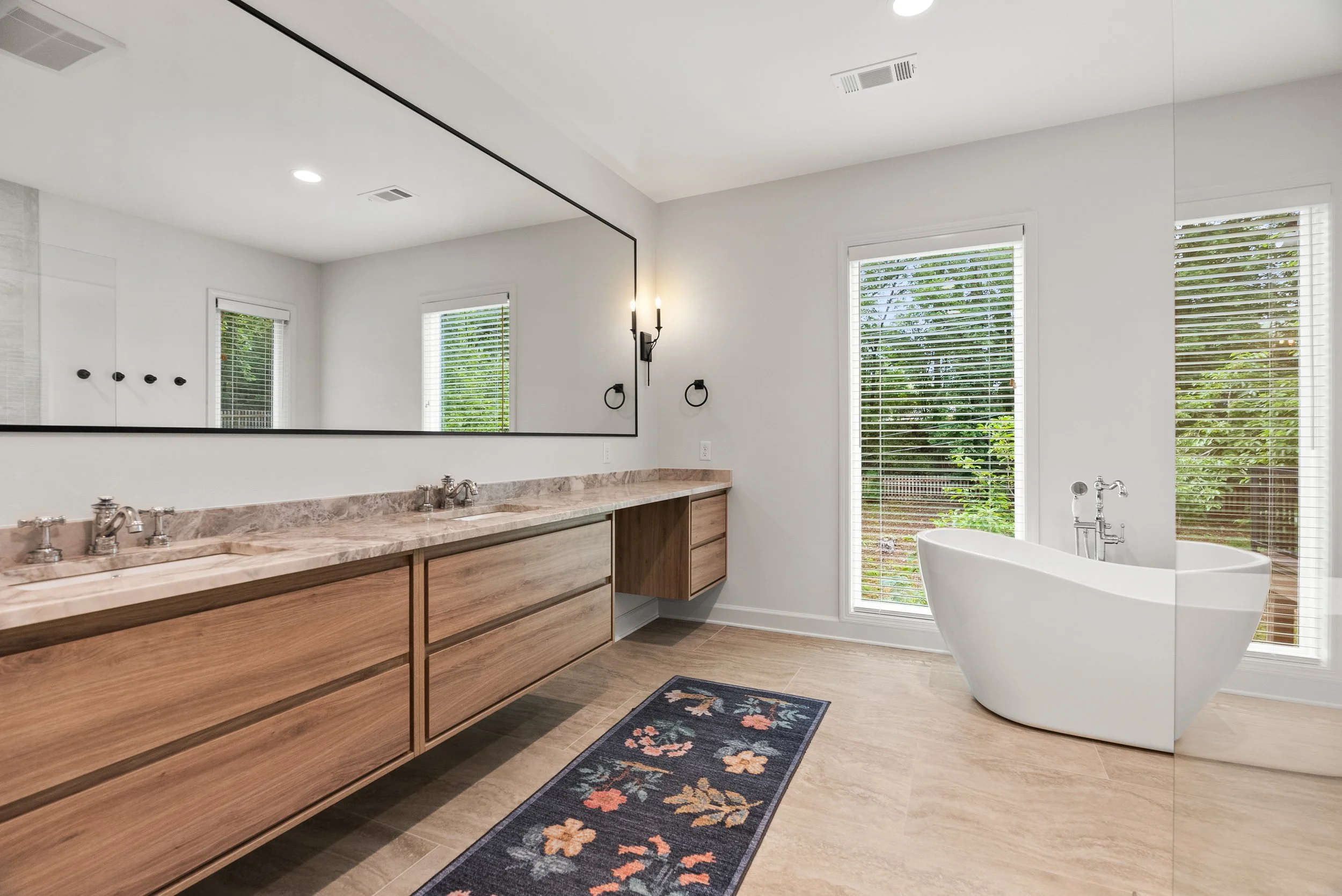 Modern bathroom with a large mirror, wooden vanity with double sinks, a white freestanding bathtub, and windows with blinds showing greenery outside.