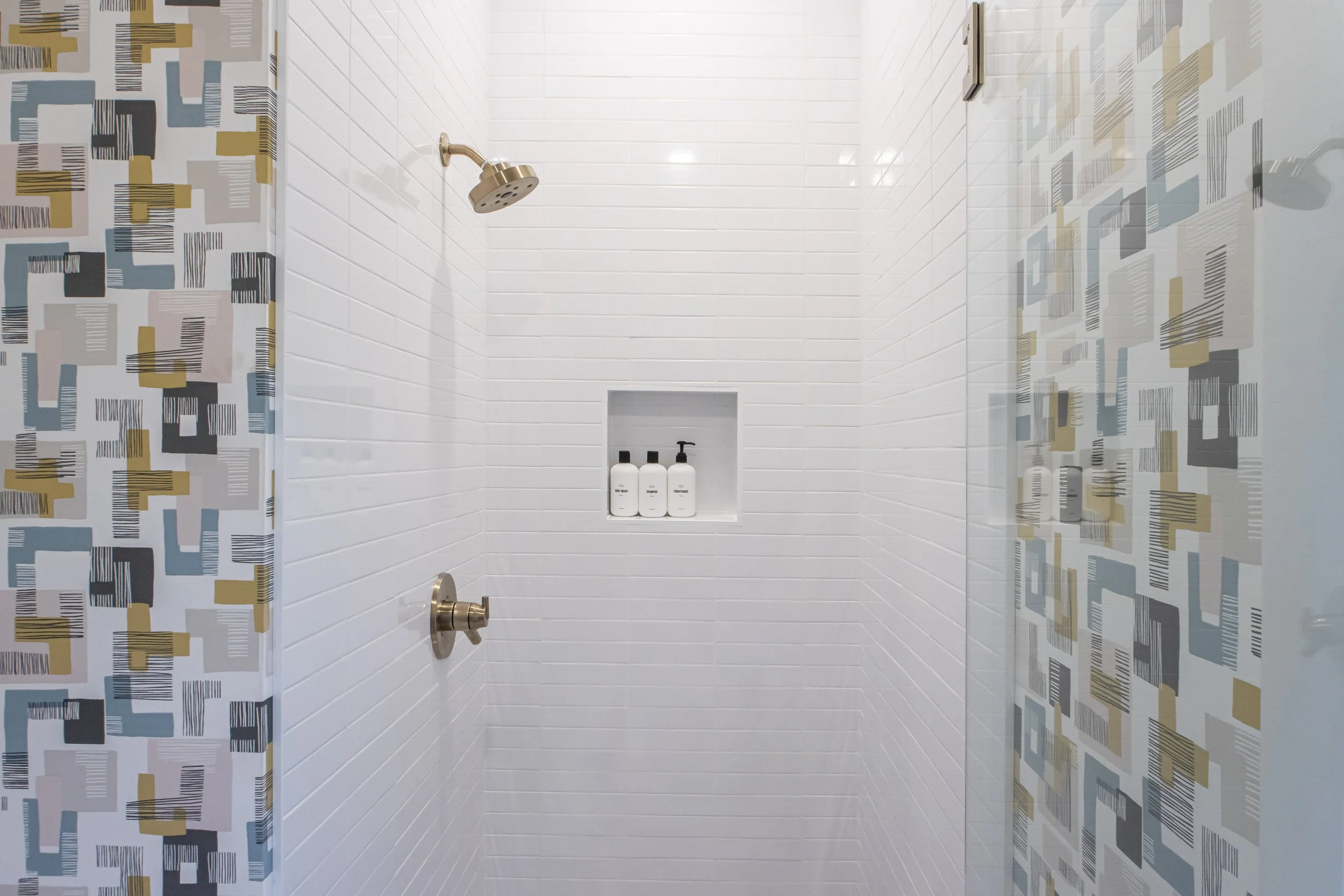 A modern shower with white tile walls, a brass shower head, and built-in shelf holding three bottles of body wash, shampoo, and conditioner. The shower is enclosed by patterned curtains with abstract rectangular designs in mustard, black, gray, and s