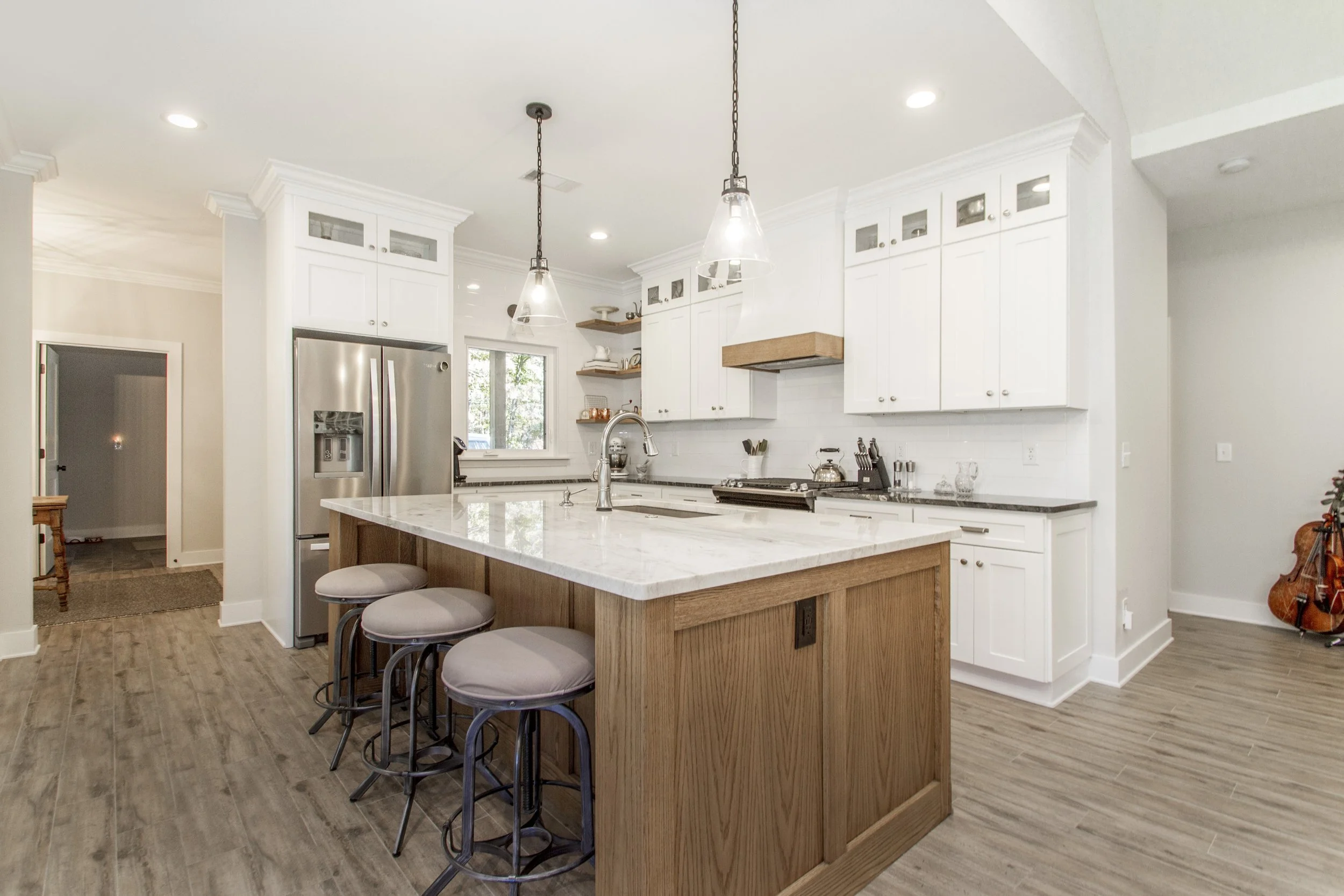 A modern kitchen with white cabinets, a marble kitchen island with wooden base, three bar stools with cushioned seats, stainless steel refrigerator, and pendant lights hanging from the ceiling.