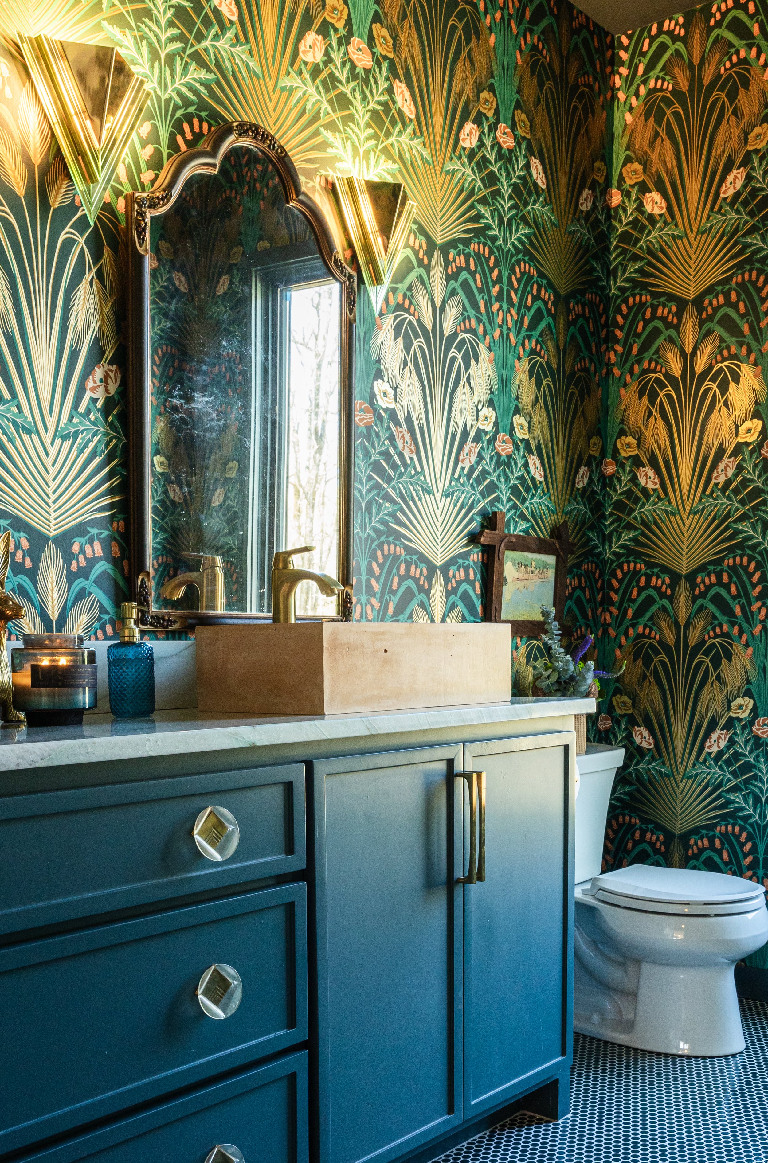 A bathroom with a bathroom vanity, a mirror with ornate details, and a toilet. The wall has a vibrant, leaf-patterned wallpaper in shades of green, gold, and coral. On the vanity, there are soap dispensers and a small decorative item. The room has a 