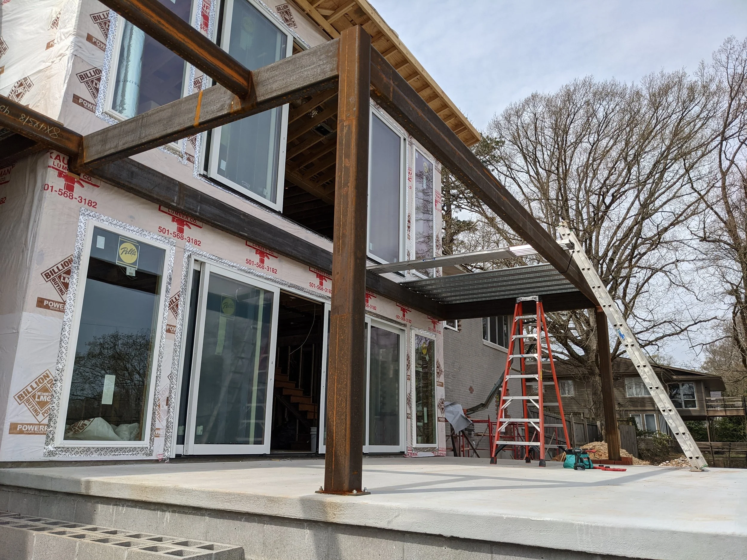 Construction site of a house with large windows, metal beams, scaffolding, and ladders outside. There are trees in the background and building materials nearby.