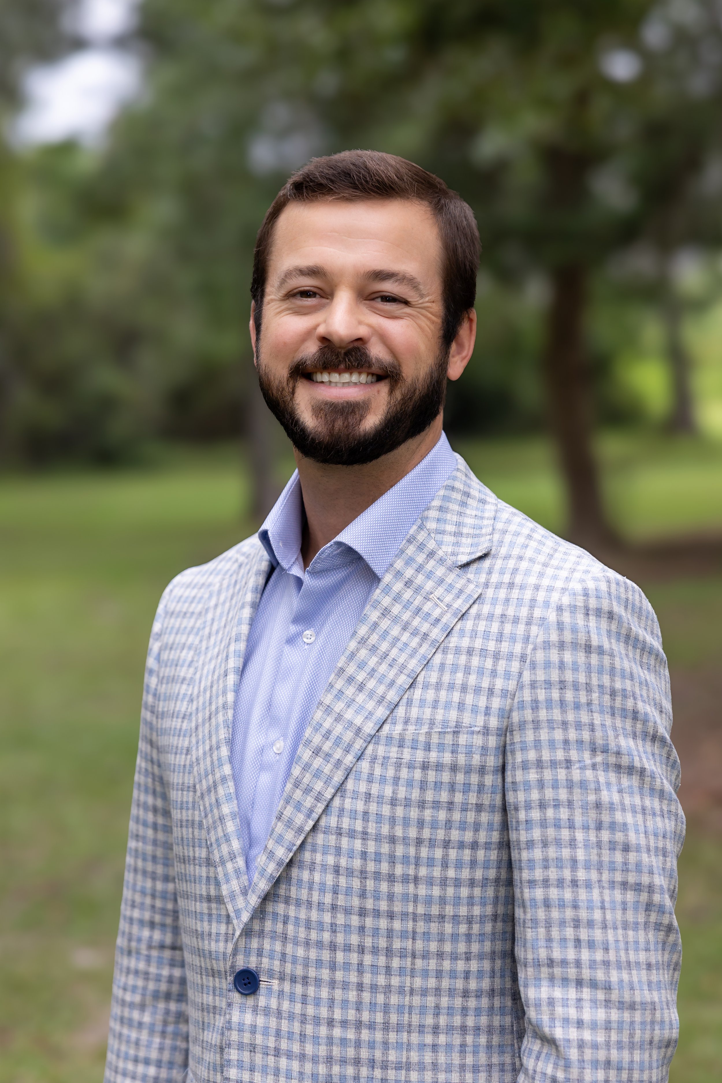 A man with a beard and short brown hair smiling outdoors, wearing a light blue dress shirt and a gray checkered blazer, with a blurred green park background.