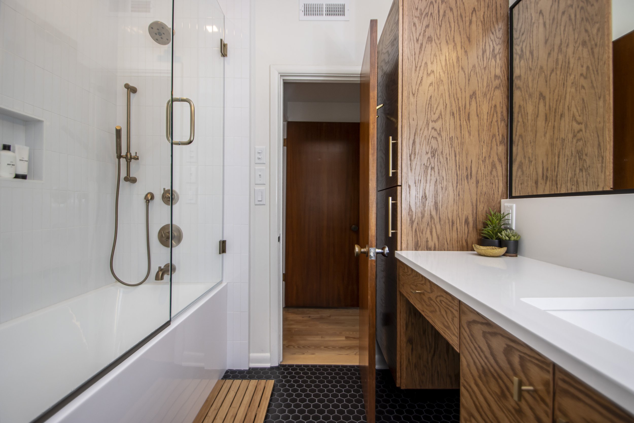 The image shows a modern bathroom with a glass shower enclosure on the left, white tiled walls, and wooden cabinets with gold handles on the right. There is a small plant on the countertop.
