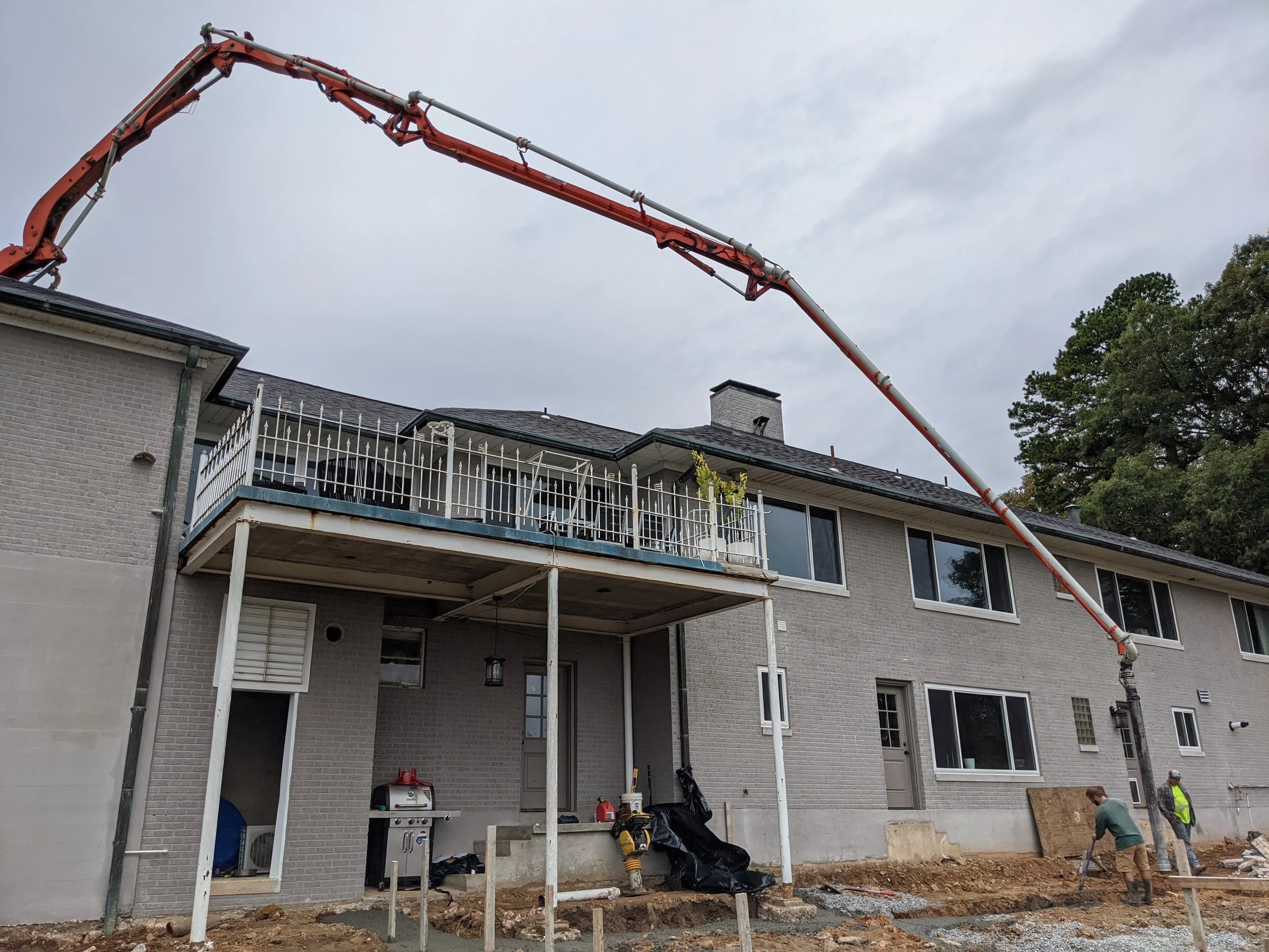 Construction workers working on an outdoor patio or deck area of a house, with scaffolding and tools, on a cloudy day.