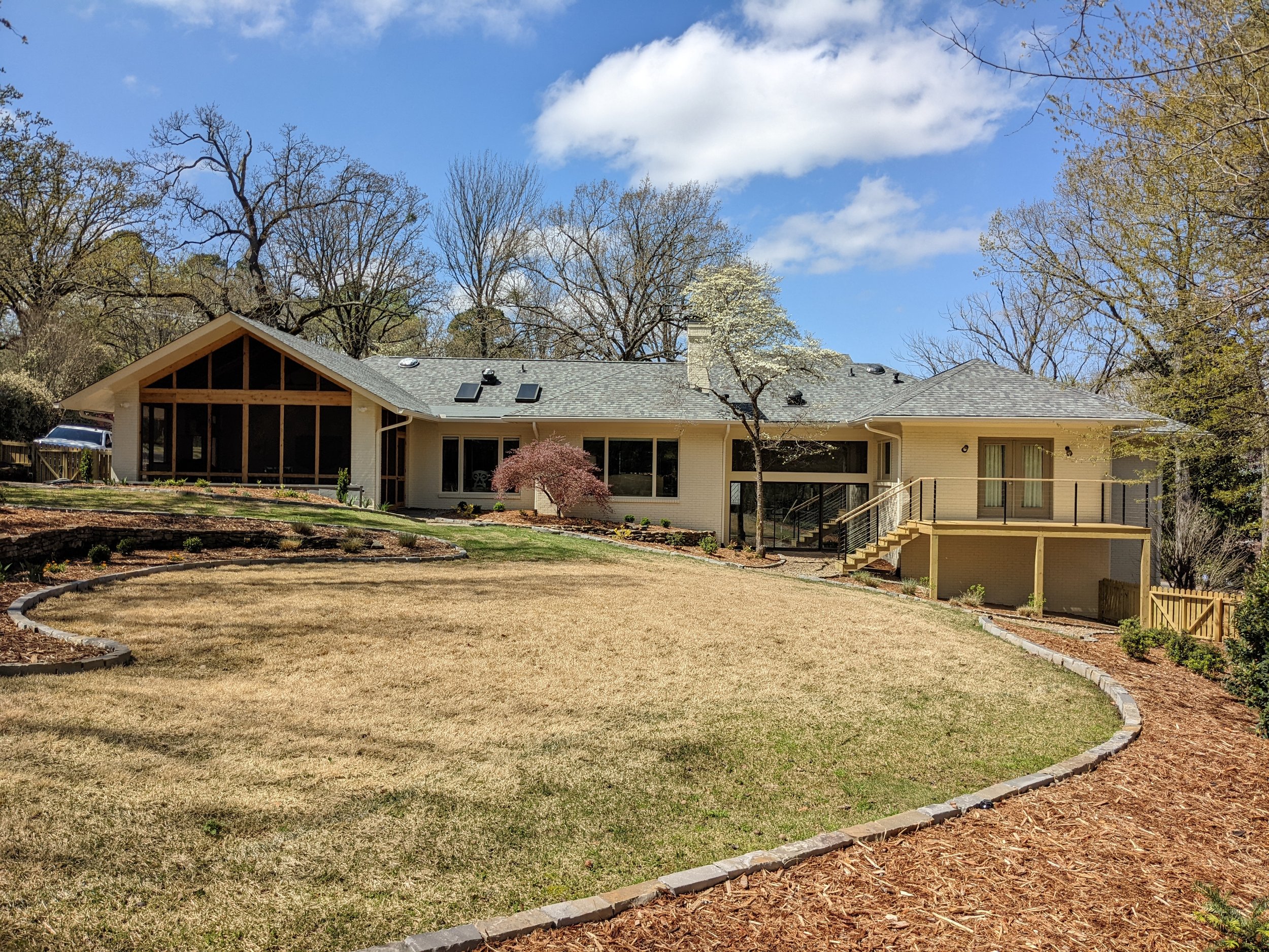 A house with a screened porch, surrounded by trees and a landscaped garden with patches of grass and mulched areas under a partly cloudy sky.