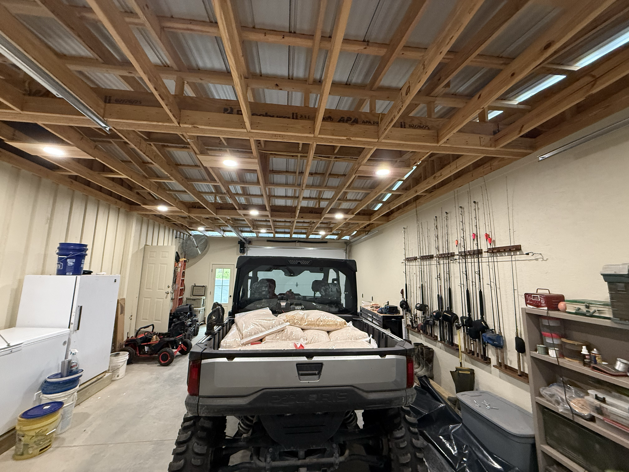 View of a garage with a truck loaded with bags, surrounded by various tools, equipment, and storage items. Wood framing on the ceiling, low lighting, and a partially visible garage door at the back.