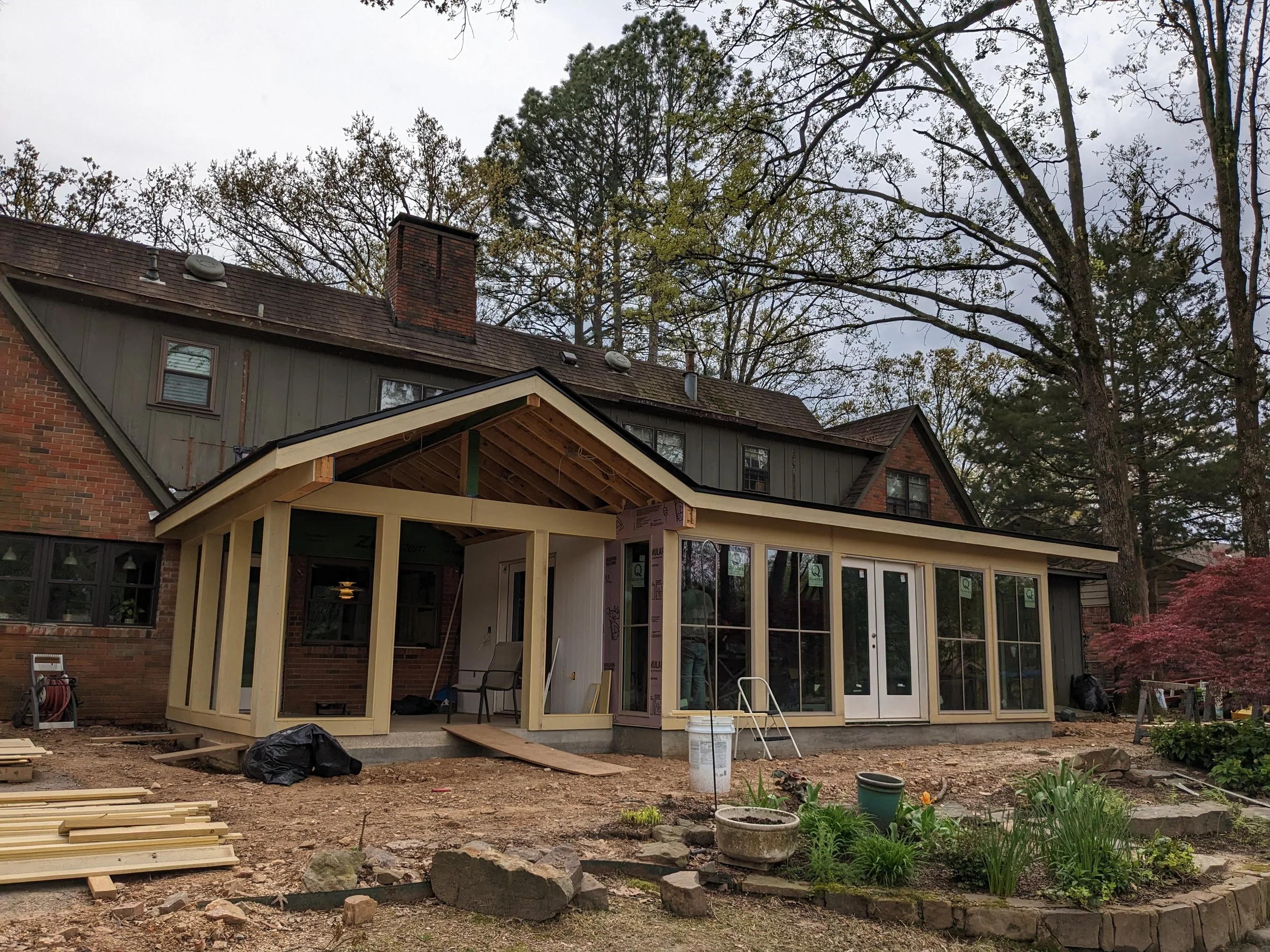 The image shows a house with a portion of the backyard under renovation. The house has a brick and gray exterior with an assortment of windows and chimneys on the roof. In the foreground, there is a construction area with wooden framing, construction