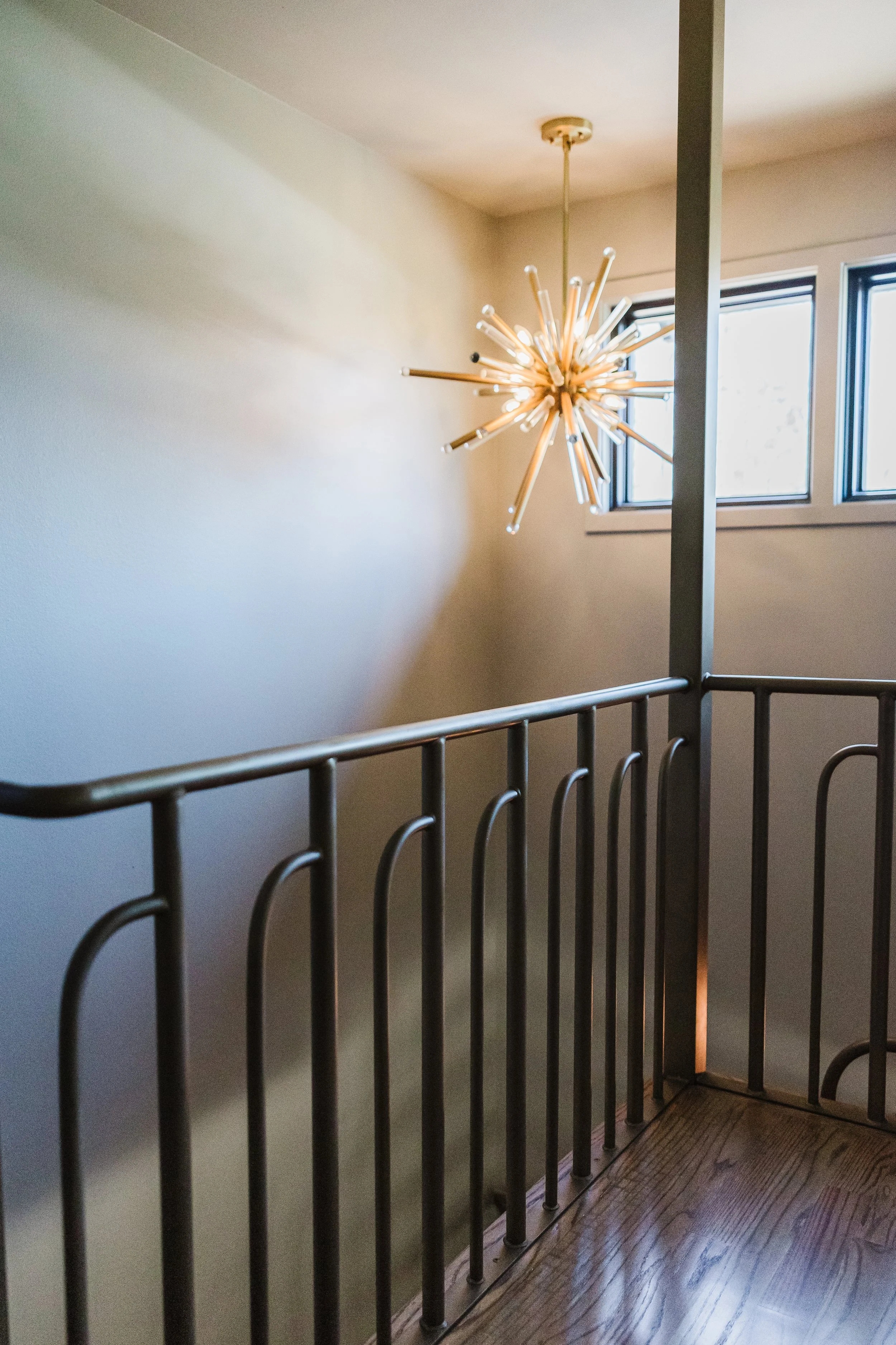 Indoor scene with natural light coming from windows, featuring a modern starburst chandelier hanging from the ceiling, and a staircase with a black metal railing. The floor is wooden with a dark finish.