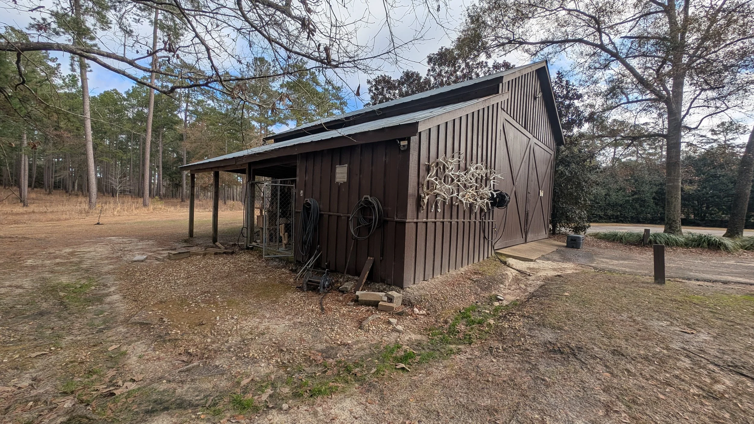 A small wooden shed painted brown with a sloped metal roof, measuring tapes, hoses, and a metal gate attached outside, surrounded by trees and an open yard.