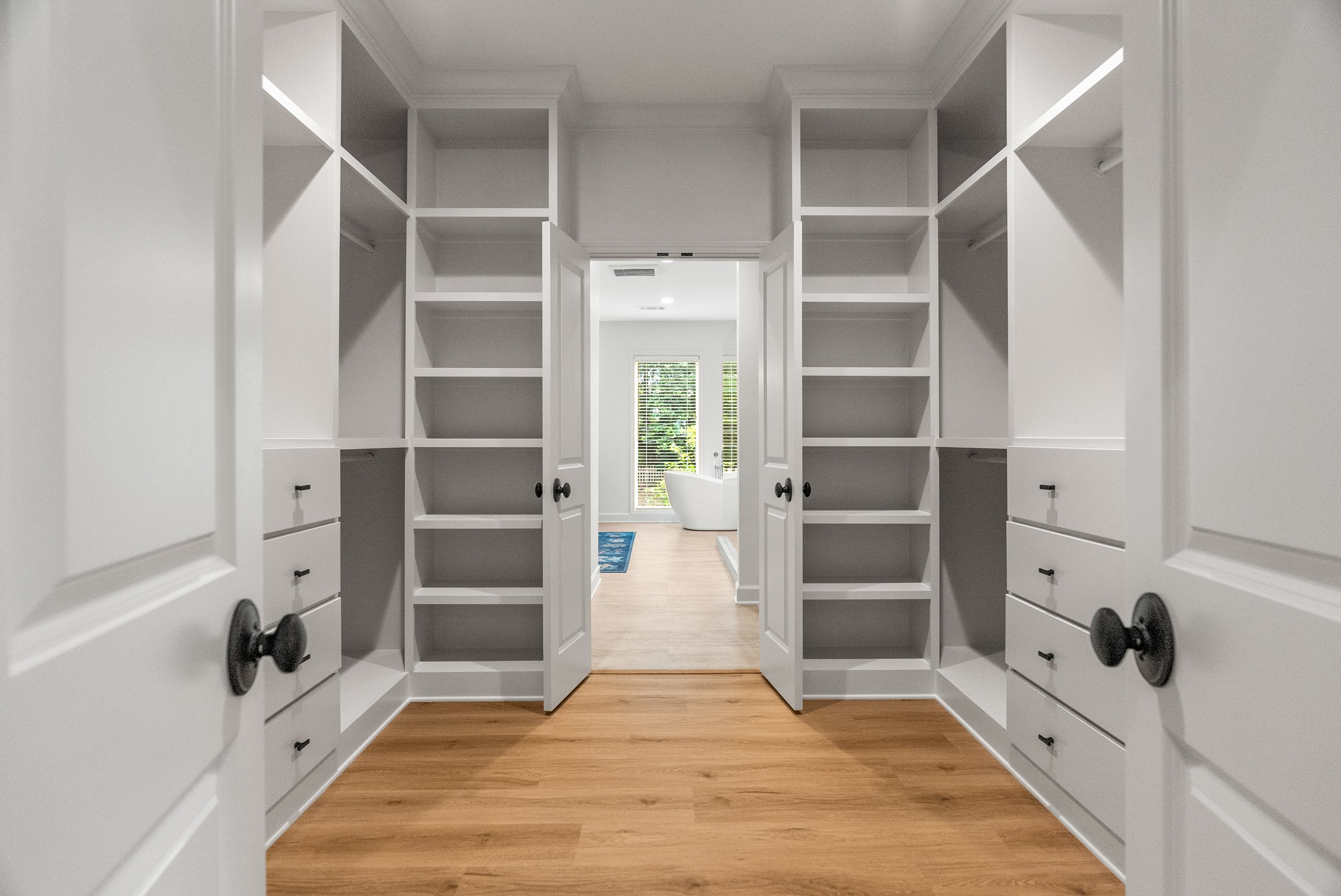 View of a walk-in closet with white built-in shelves and drawers, open double doors, hardwood floor, and a doorway leading to a bathroom with a bathtub and window.