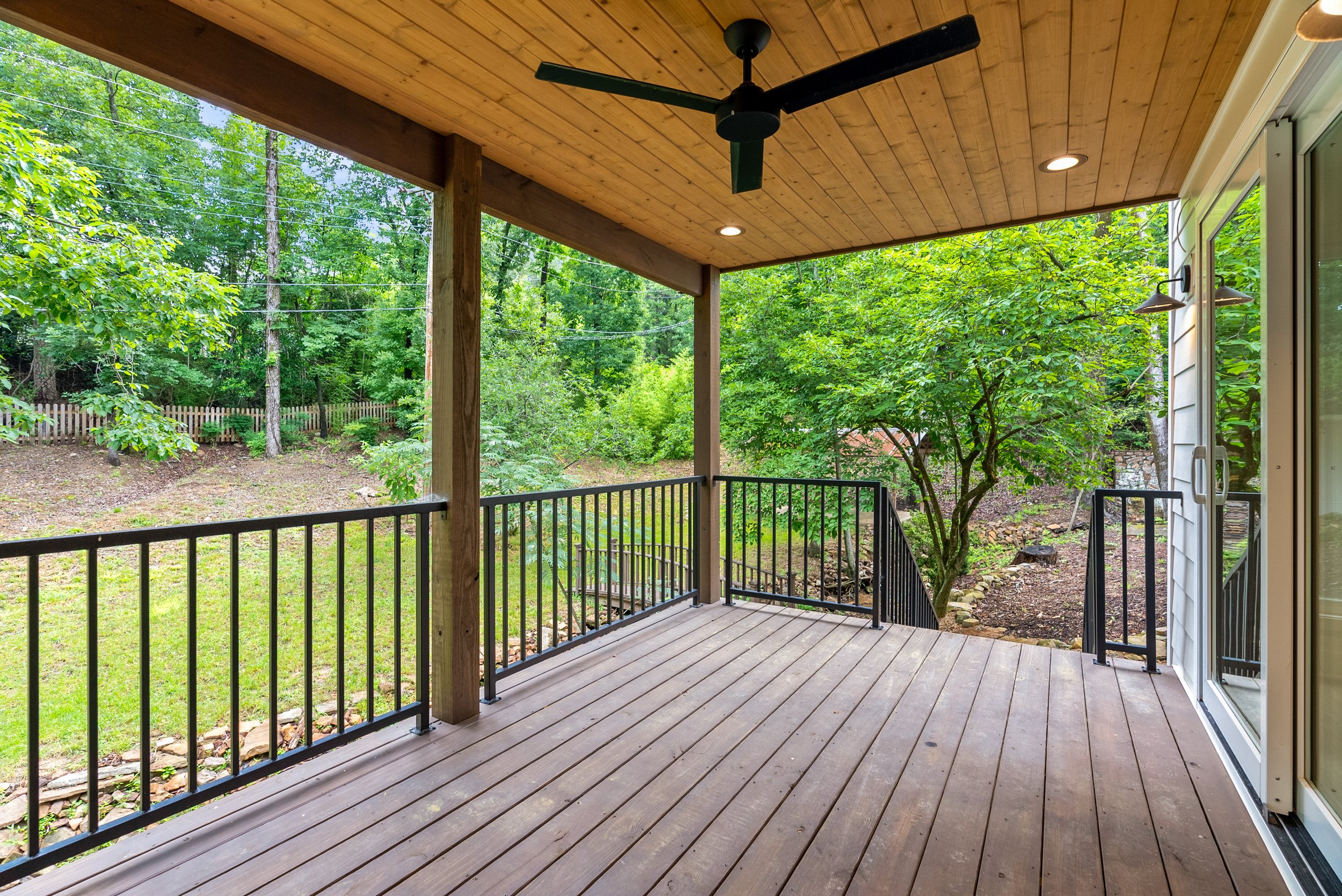 Wooden porch with black railing, ceiling fan, and recessed lighting, overlooking a lush green backyard with trees and a wooden fence.