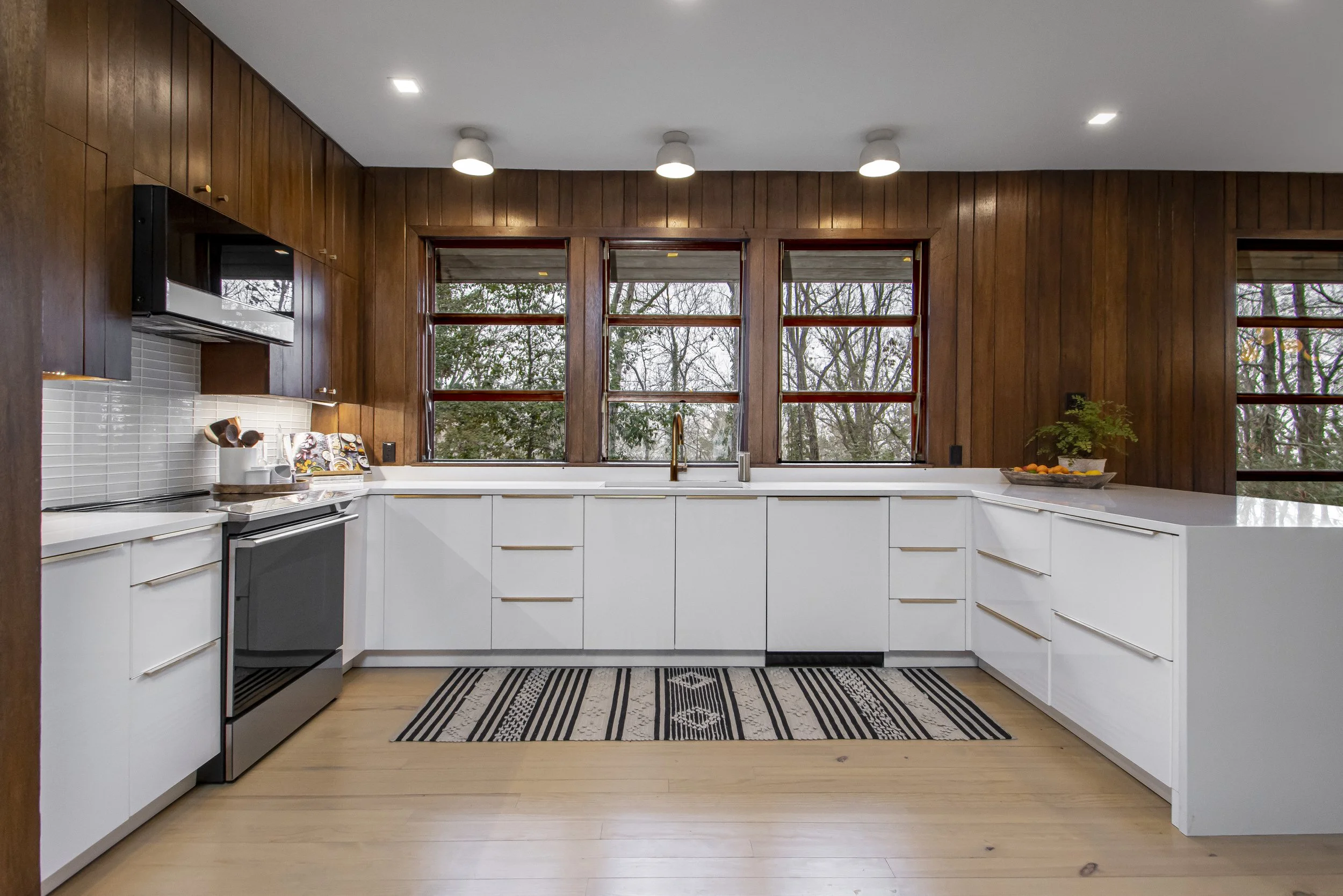 Modern kitchen with white cabinetry, a black oven, a white countertop, wood-paneled walls, large windows showing trees outside, and a black and white striped rug.