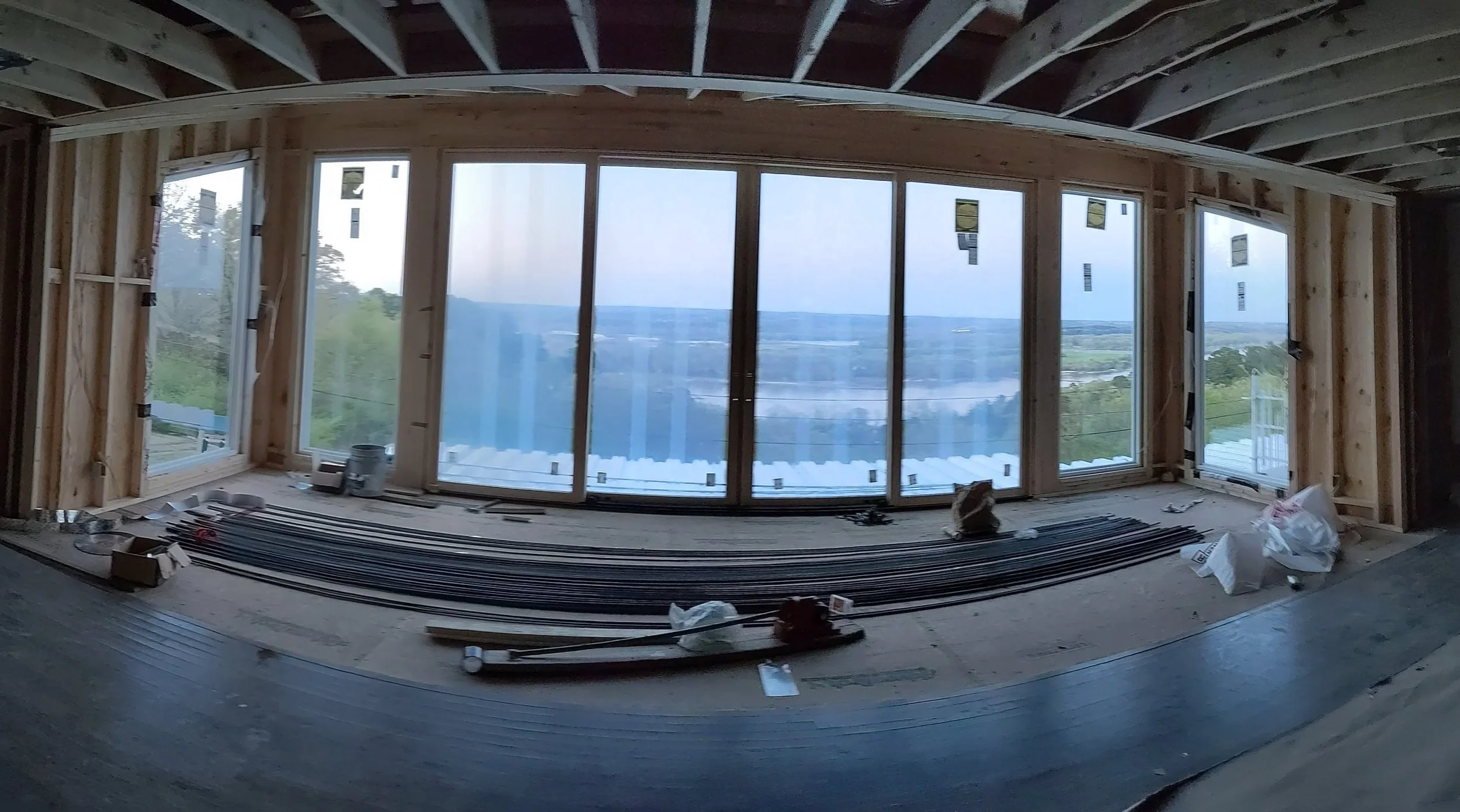 Interior of a building under construction with large windows overlooking a landscape, exposed wooden beams, construction tools, and materials scattered on the floor.