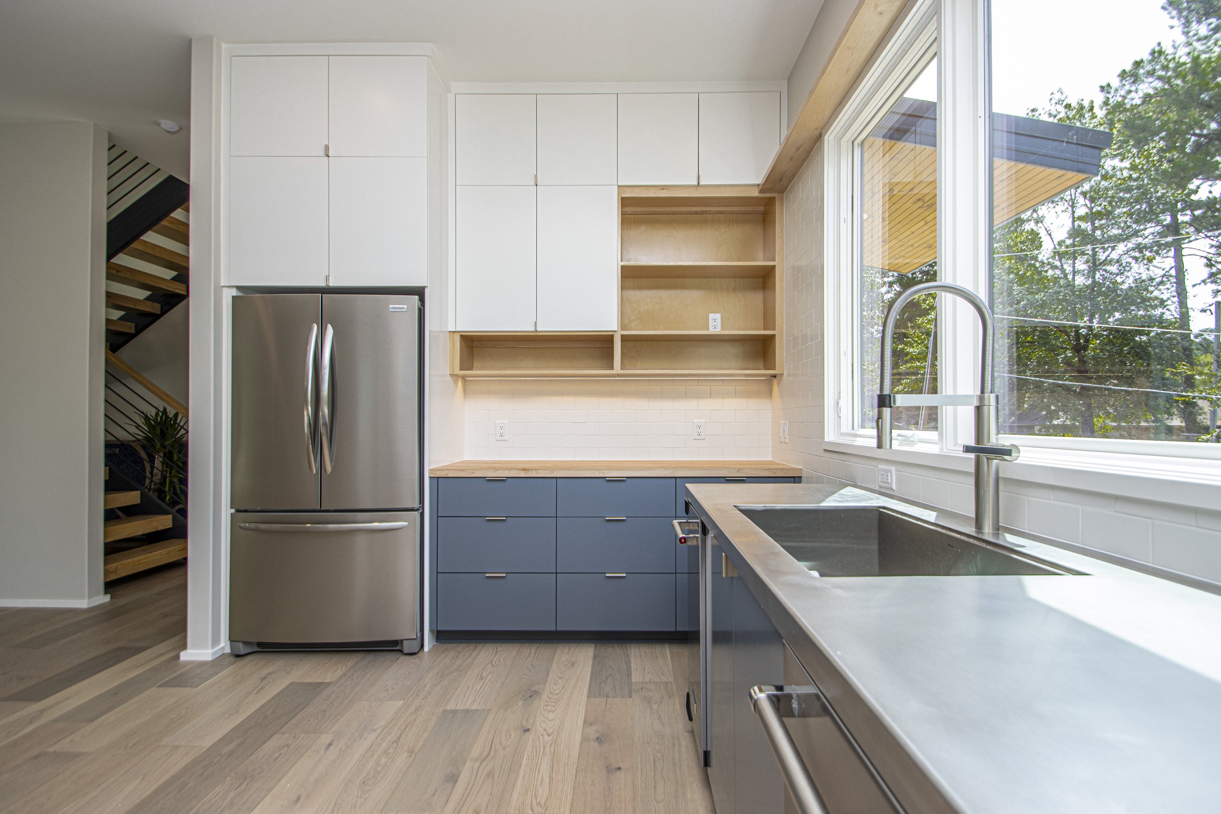 Modern kitchen with wooden cabinetry, stainless steel refrigerator, large window, and wooden flooring.