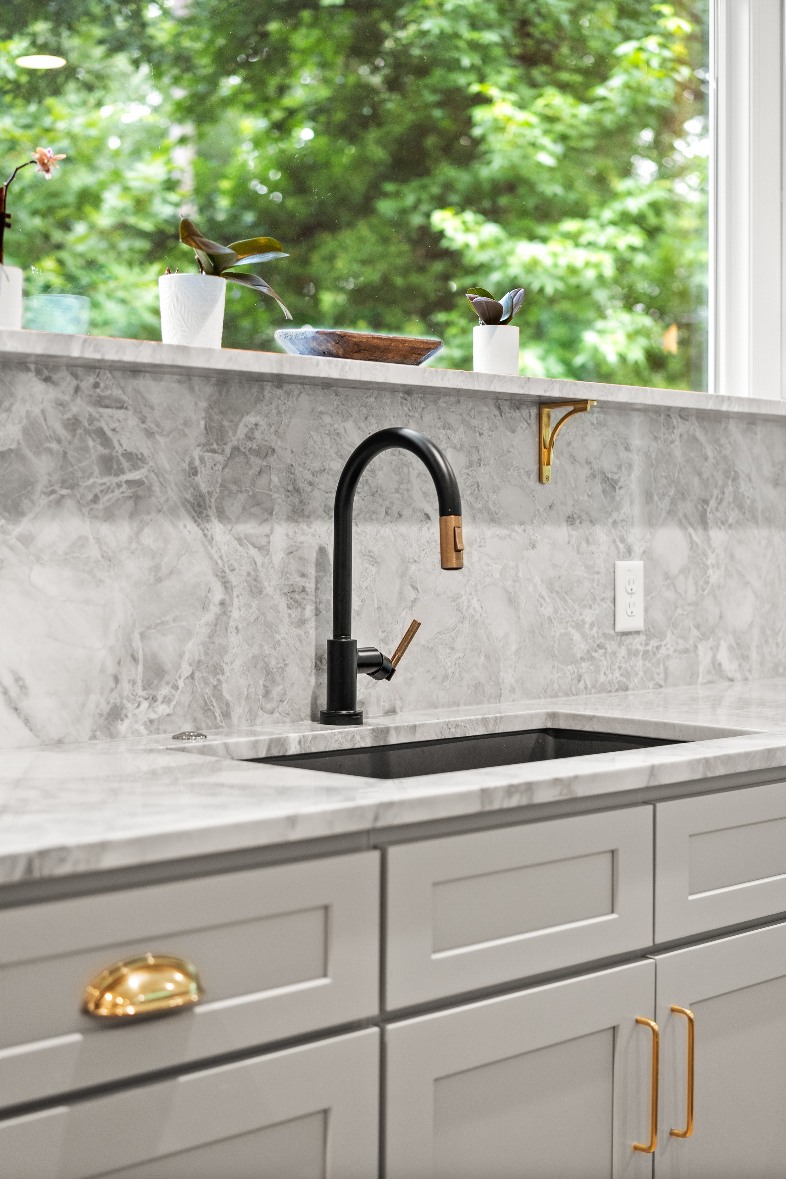 A kitchen countertop with a marble surface, a black faucet, white cabinets with gold handles, and a window with green trees in the background. Decor items on the windowsill include a potted plant, a wooden bowl, and a small ceramic vase.