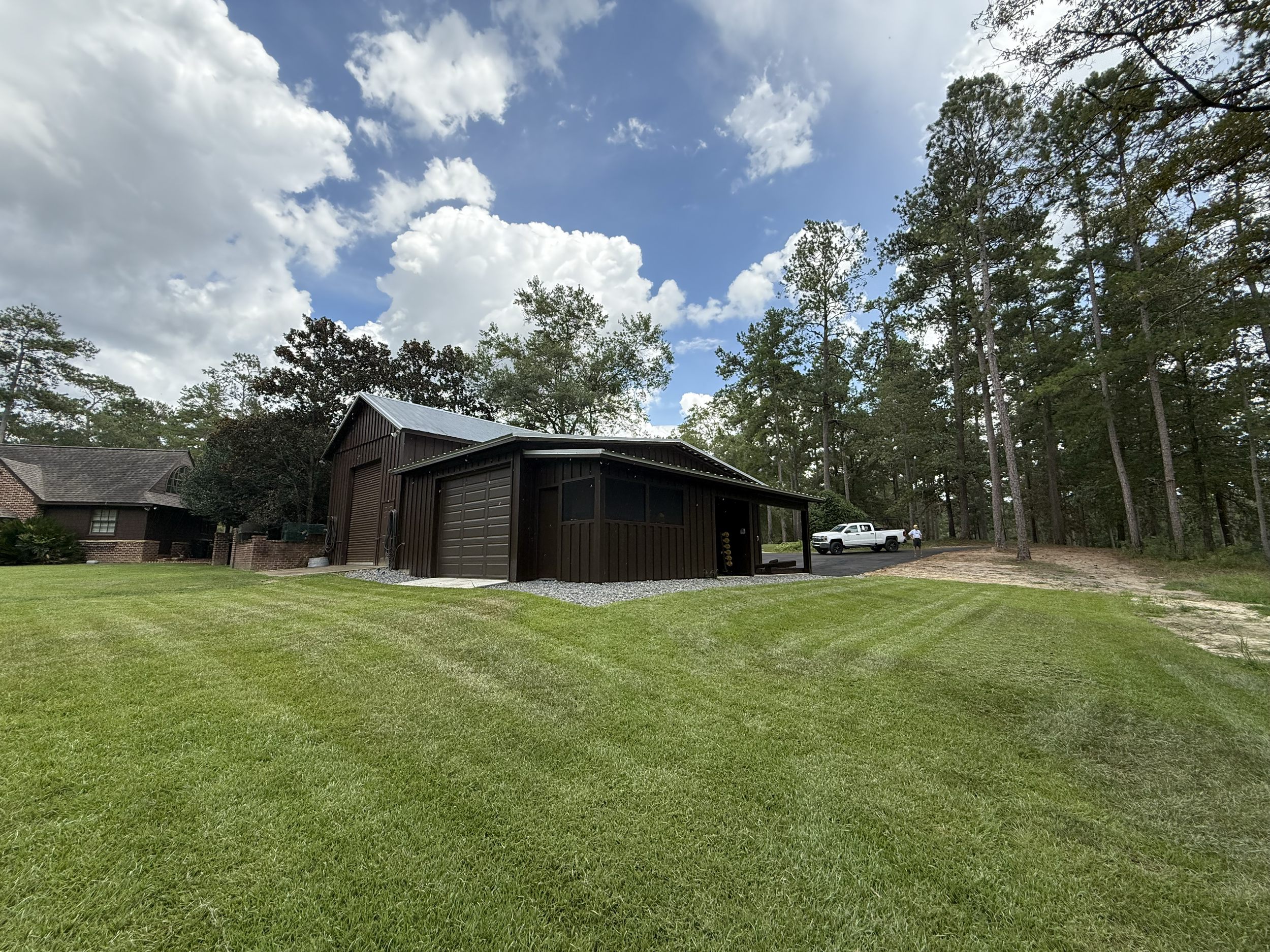 A residential backyard with a three-car garage, a lawn, tall trees, a white pickup truck, and a person walking in the distance under cloudy skies.