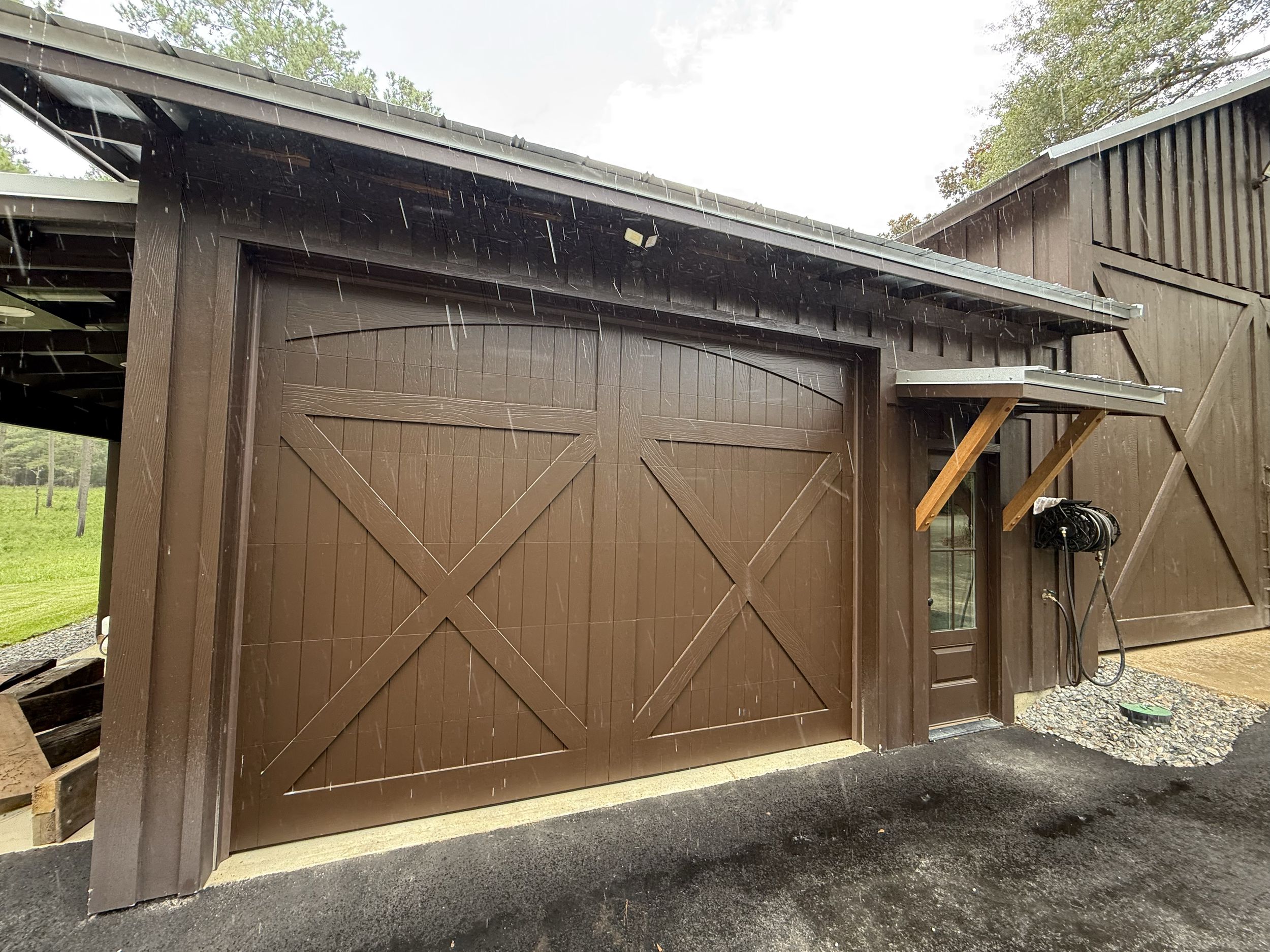 A brown wooden barn with a garage door, rain falling onto the roof, and a large tree in the background.