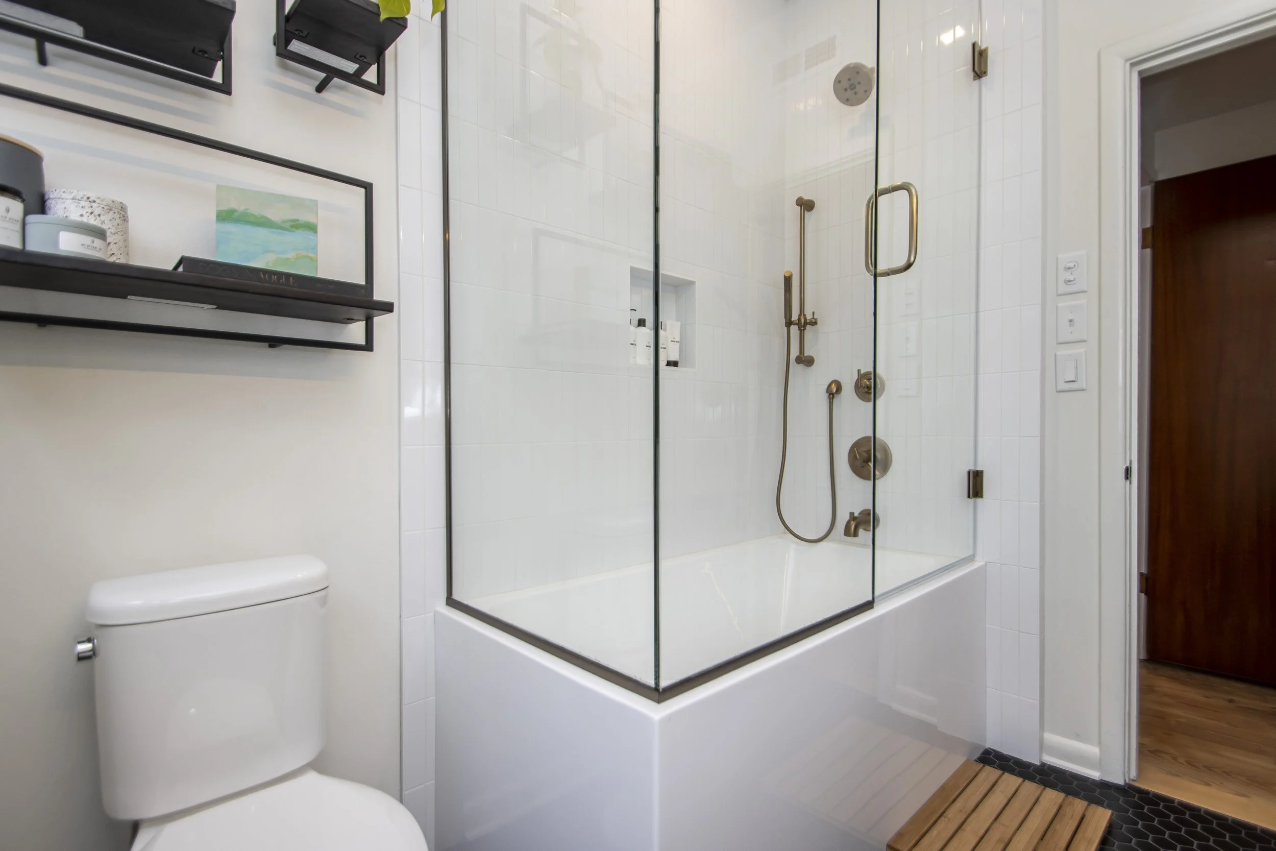 Bathroom with a glass-enclosed shower featuring brass fixtures and a wooden bath mat outside the shower. The wall is white tiled, and there is a white toilet partially visible on the left. The right side has a doorway leading to a room with wooden fl