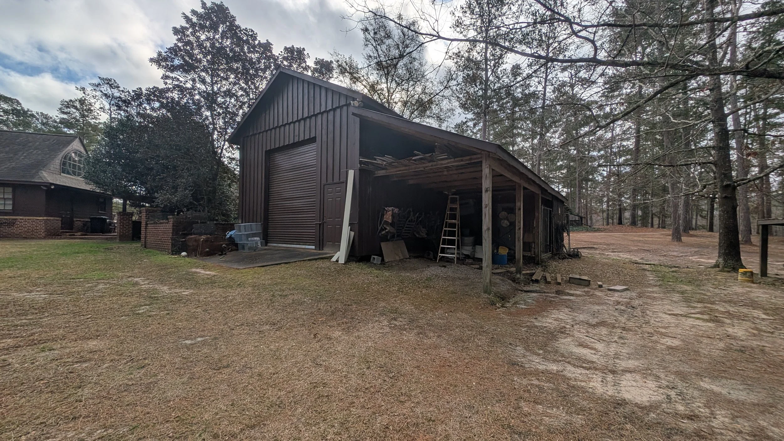 A rural backyard scene showing a wooden shed with a metal door, a brick house to the left, and trees in the background. The shed has an open side with tools and miscellaneous items inside. The ground is a mix of dirt and grass.