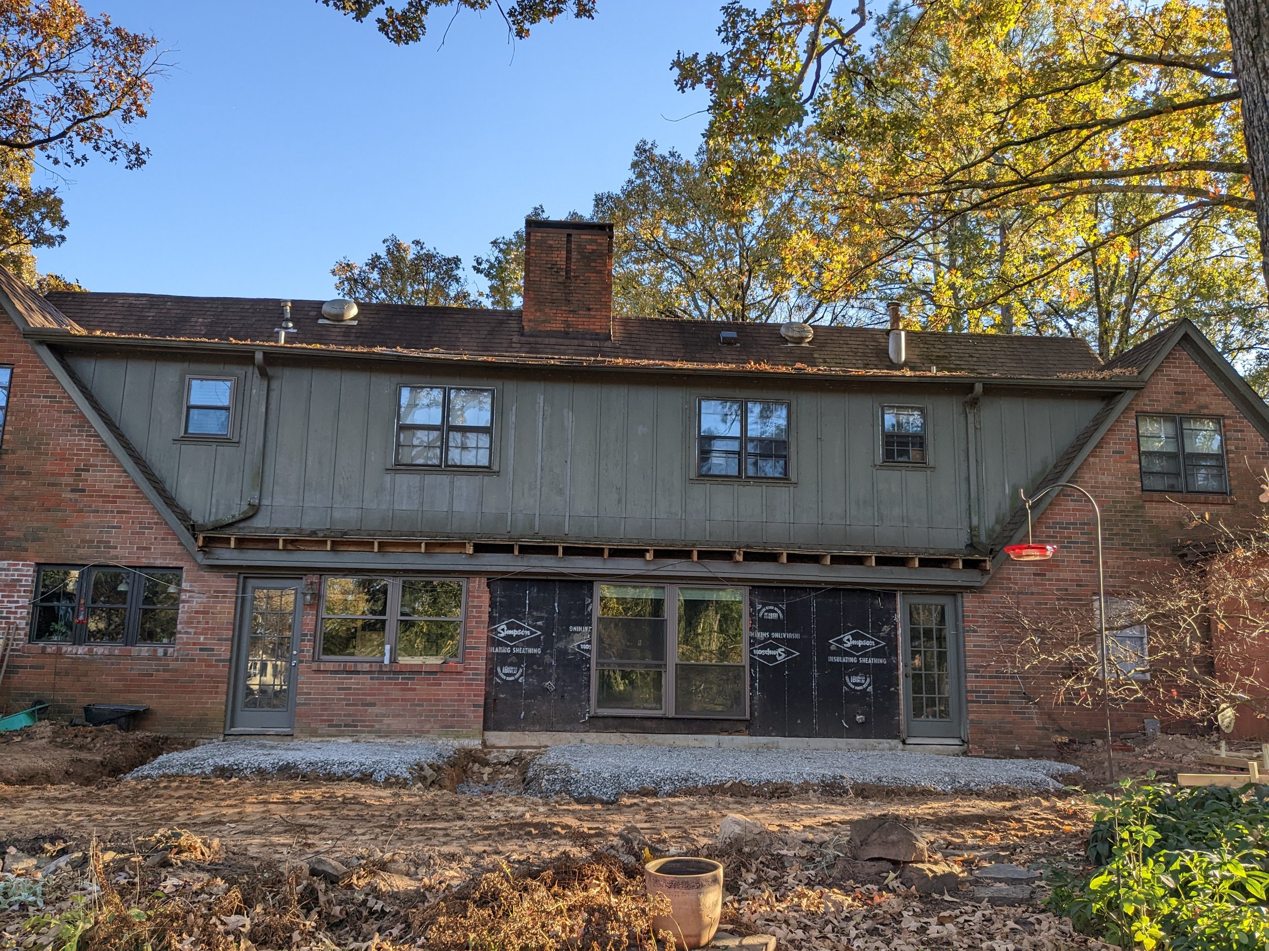 This is a photo of the back of a house under construction or renovation with some ground work done, showing a partially finished patio or walkway area, with trees and a clear blue sky in the background.