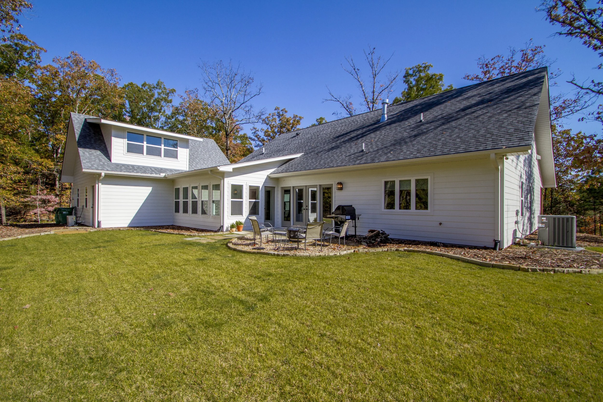 White house with large backyard and patio furniture, surrounded by trees with autumn leaves, under a clear blue sky.