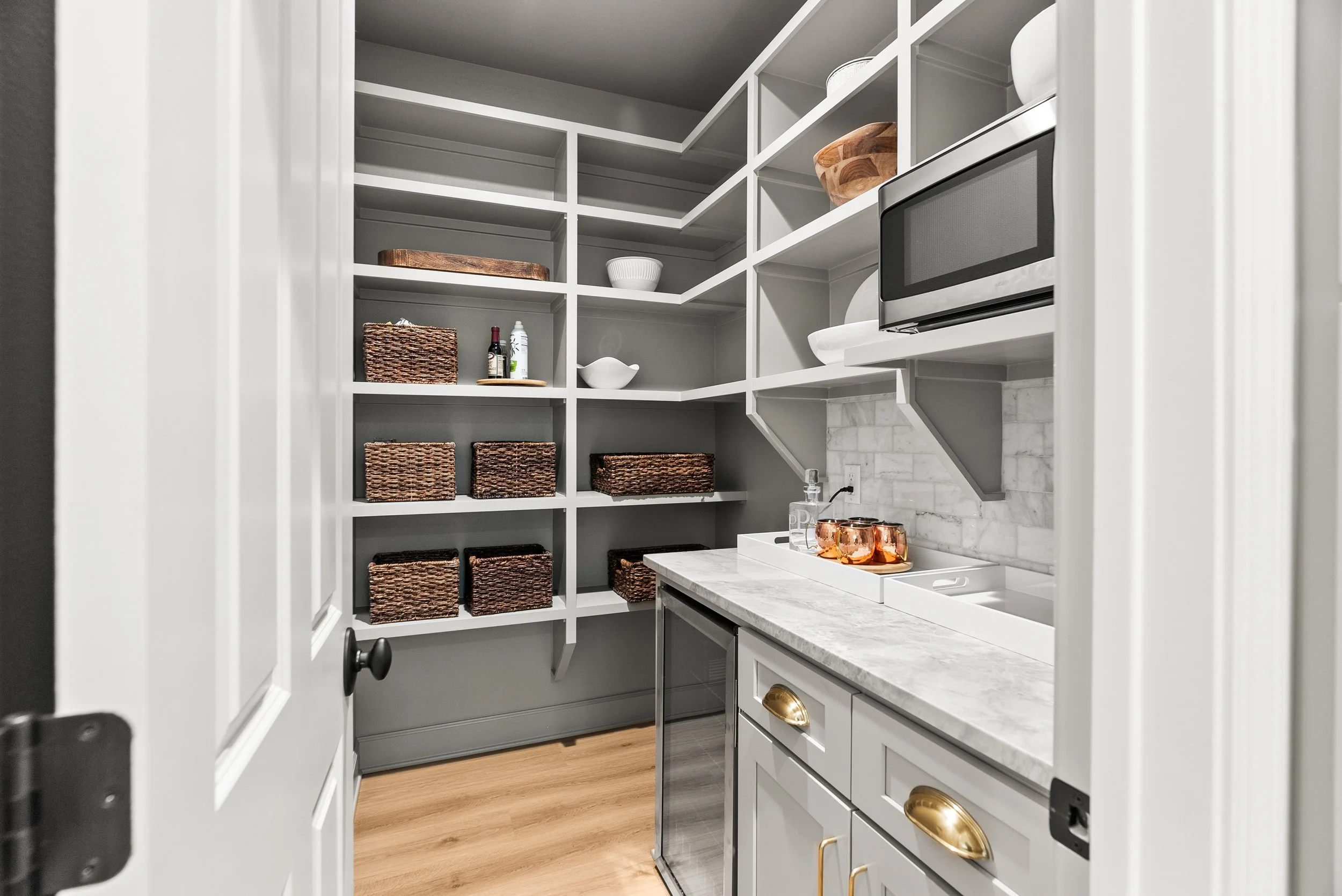 A modern pantry with white shelving, some with wicker baskets, a microwave, a marble countertop, and various small kitchen items.