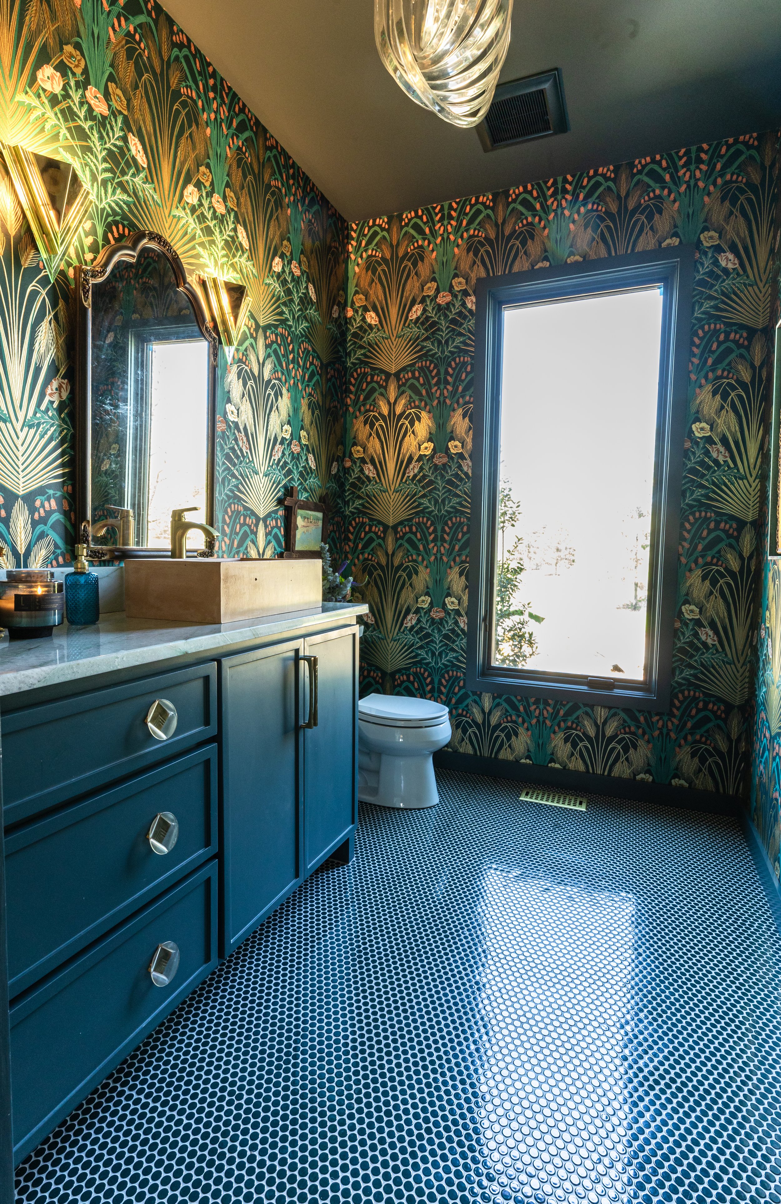 A bathroom with dark green and gold botanical wallpaper, a blue vanity with silver handles, a rectangular vessel sink, a mirror, wall-mounted light fixtures, a window letting in bright natural light, a white toilet, and a mosaic tile floor.