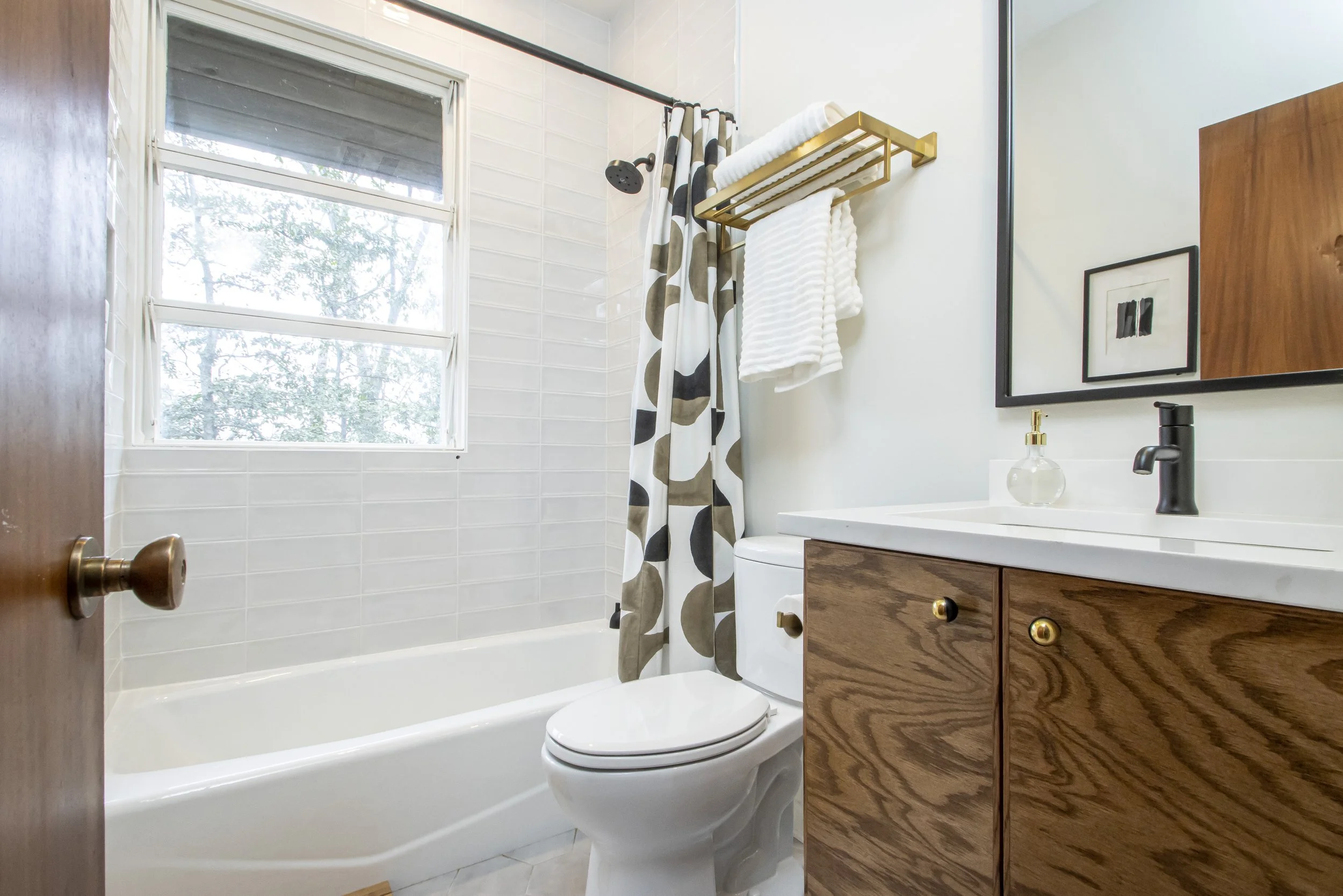 A small bathroom with a large window, a bathtub with a polka dot shower curtain, a toilet, a wooden vanity with gold knobs, a framed mirror, a soap dispenser, and a towel rack with white towels.