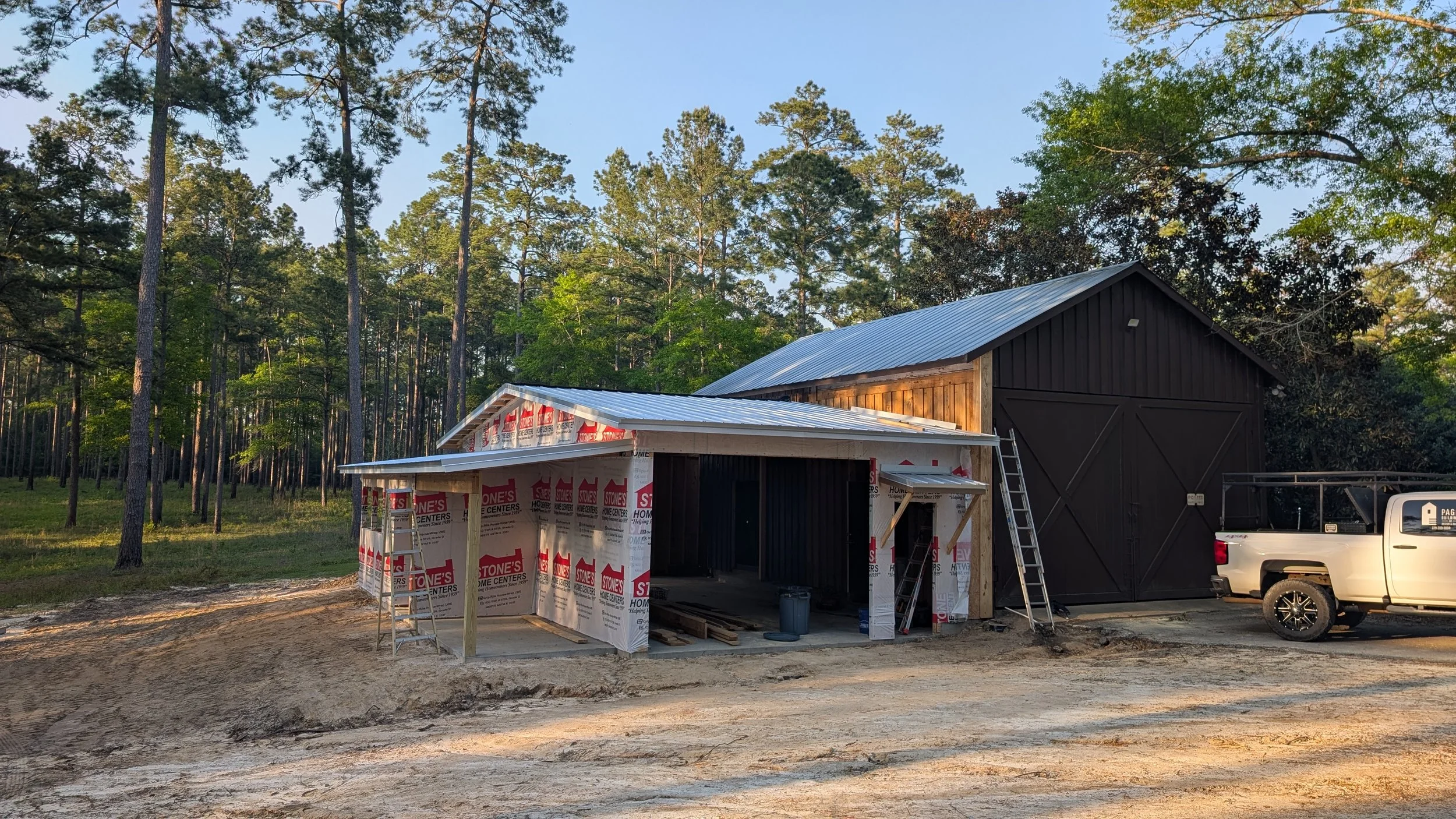 A black barn is under construction next to a wooded area, with a small extension being built covered in house wrap and a ladder leaning against it, a white pickup truck parked nearby.