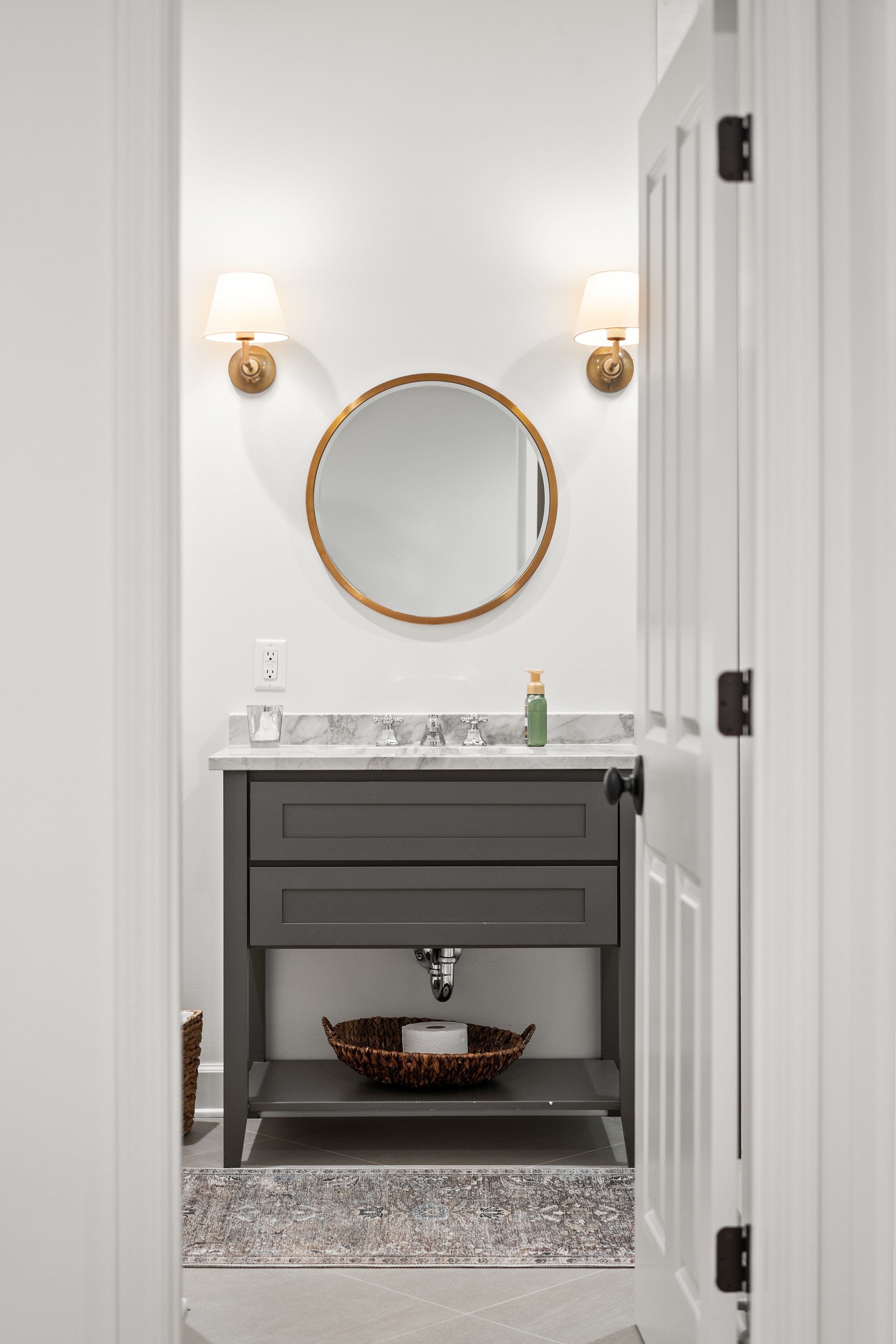 A farmhouse-style bathroom vanity with a marble countertop, gray cabinet, and a round mirror. Wall-mounted brass lamps with white shades are on either side of the mirror. Below the vanity, there is a woven basket and a rug with a vintage pattern.