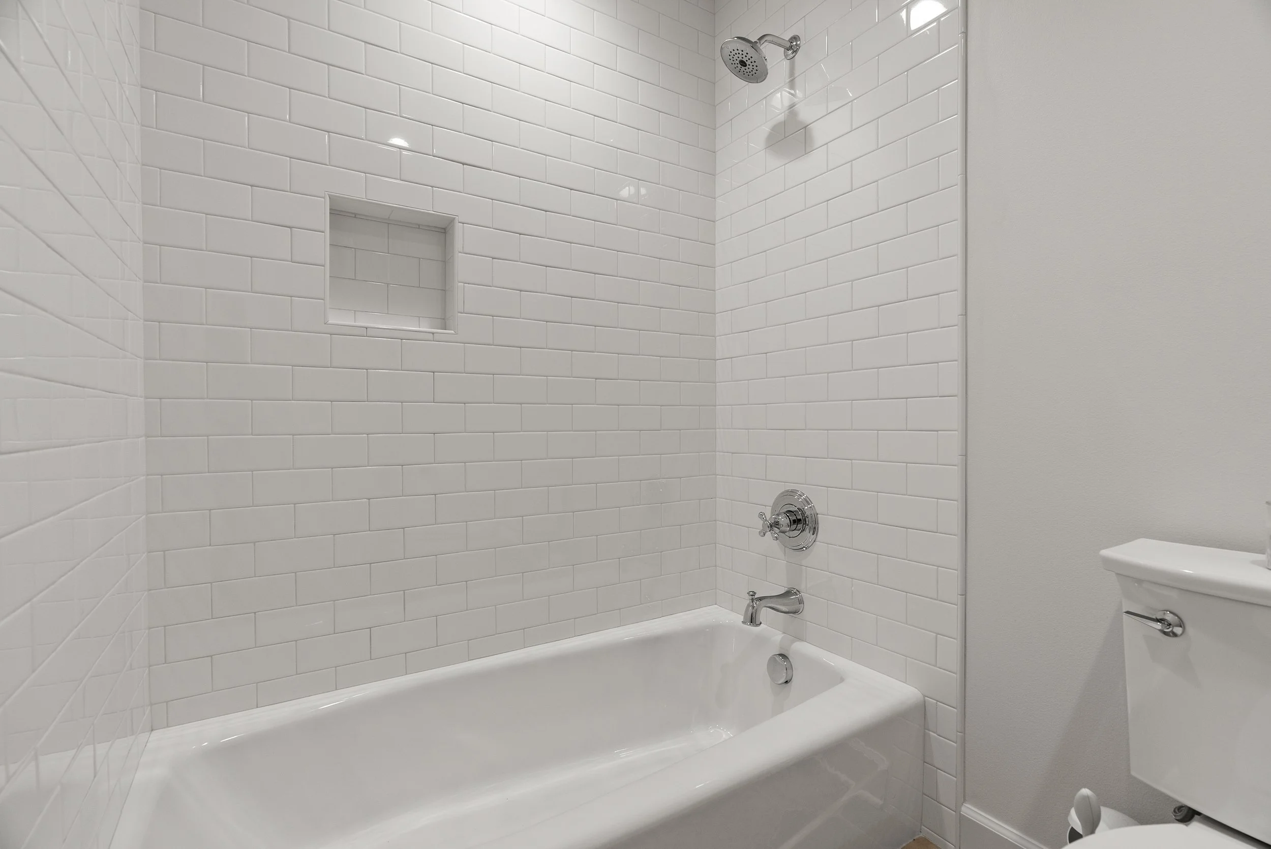 A bathroom shower with white subway tiles, a built-in soap niche, a showerhead, a bathtub faucet, and a partial view of a toilet.