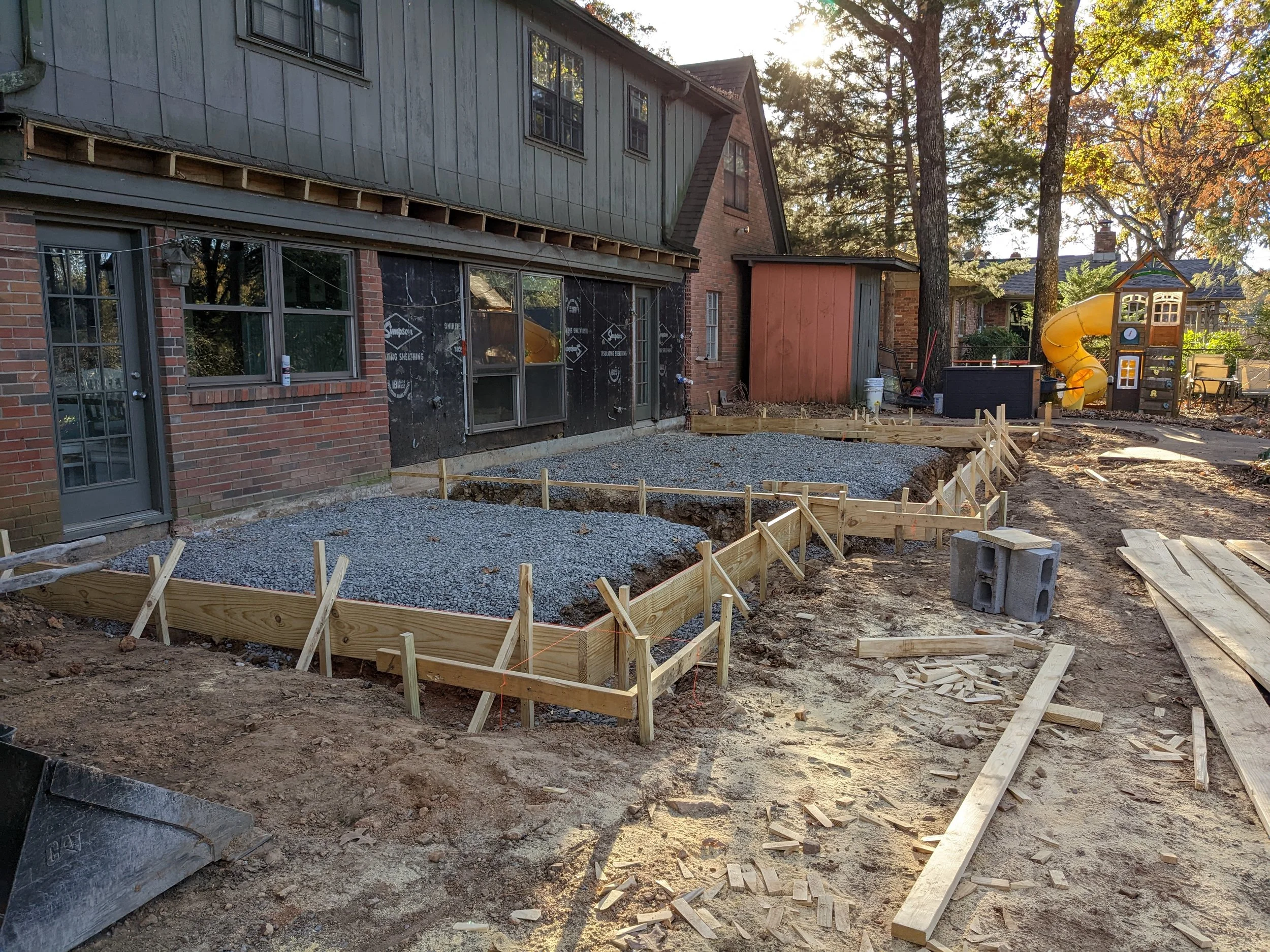 Construction site in backyard with framed garden bed, gravel area, and a wooden playhouse with a yellow slide surrounded by trees.