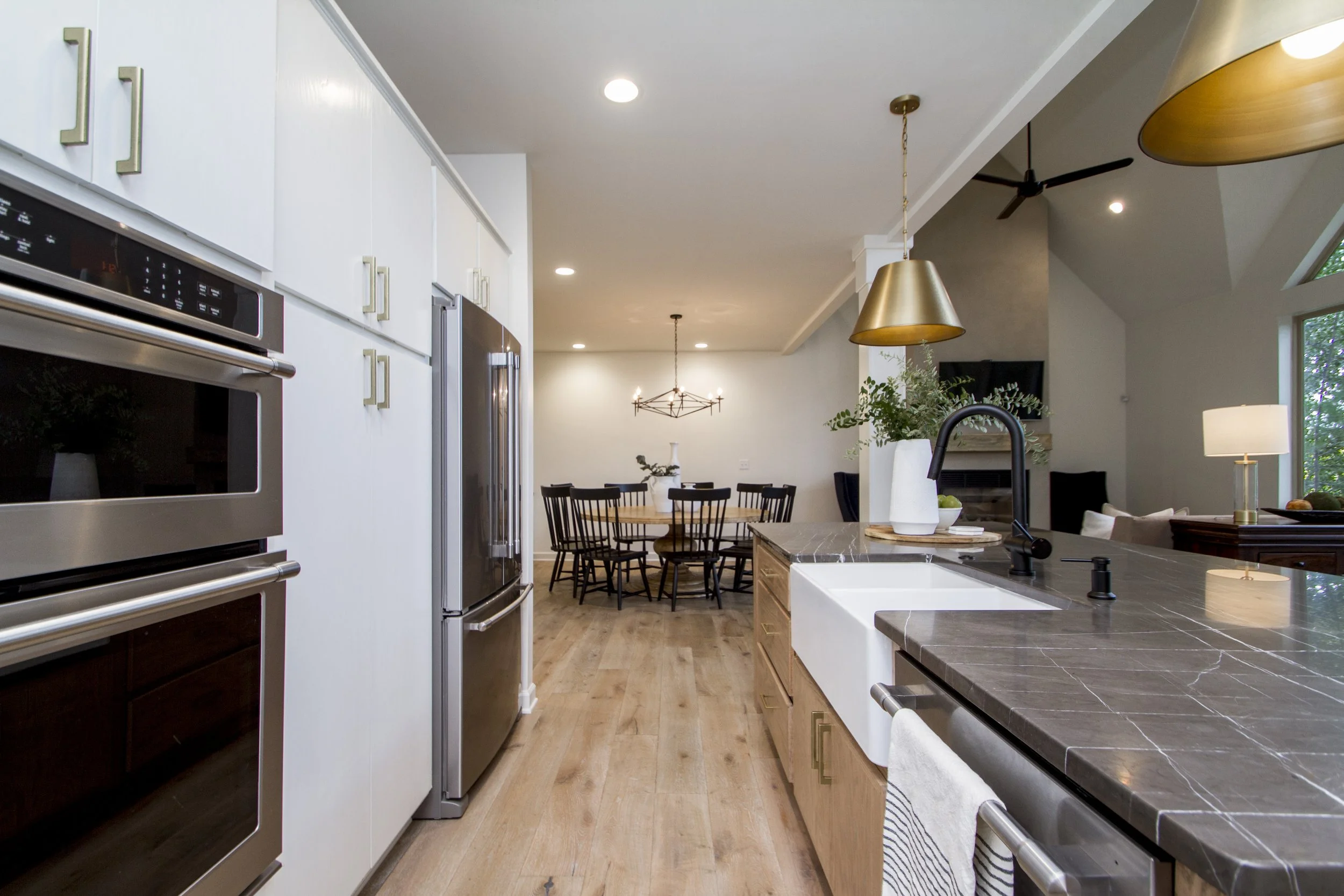 A modern open-plan kitchen and dining area with white cabinetry, stainless steel appliances, a black faucet, a marble island, and a round wooden dining table with black chairs. There are pendant and ceiling lights, large windows, and a high vaulted c