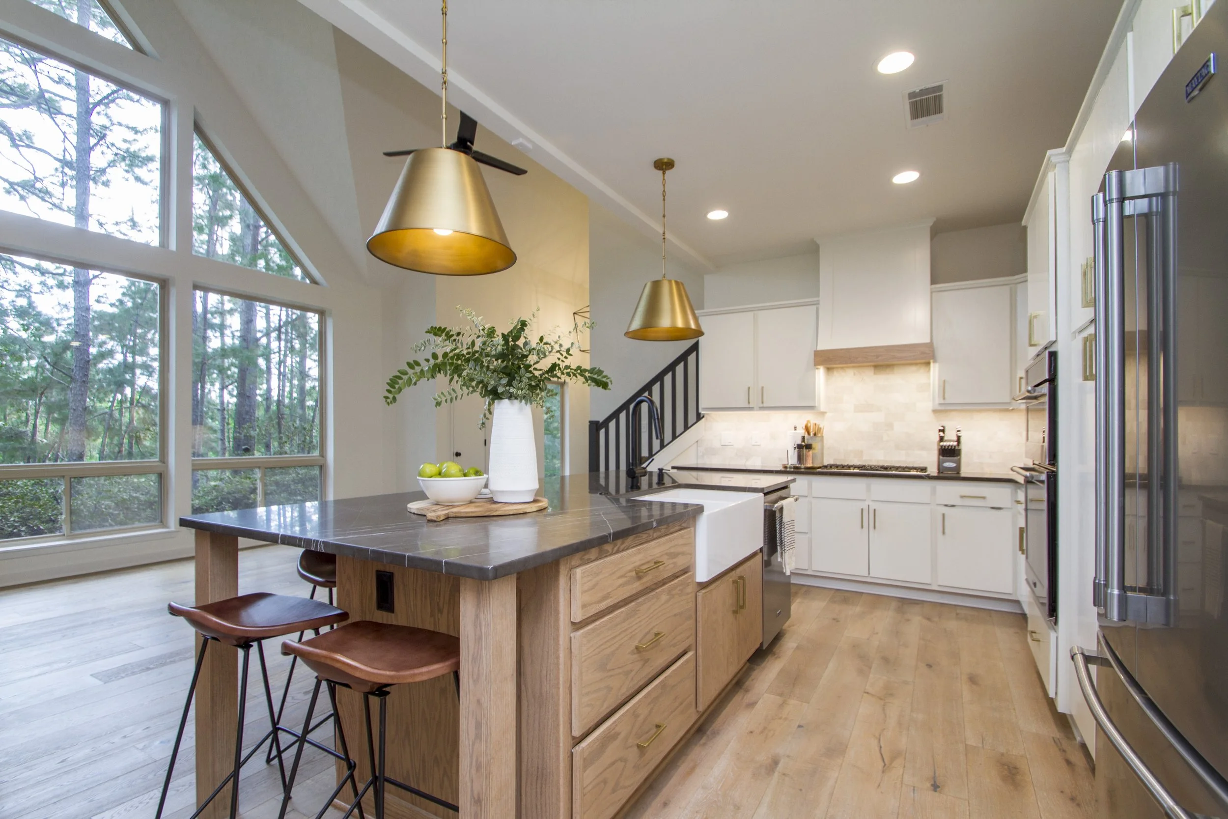 Modern kitchen with large windows, a central island with a dark countertop, wooden cabinets, gold pendant lights, and a stainless steel refrigerator. There is a tall white vase with greenery and a bowl of green apples on the island.