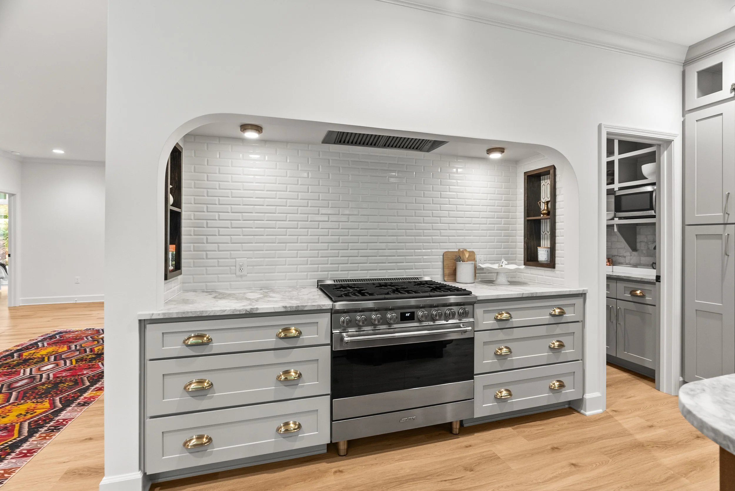 Modern kitchen with white subway tile backsplash, gray cabinets with gold handles, and a stainless steel oven and stove. White marble countertops and a colorful rug on a wooden floor.