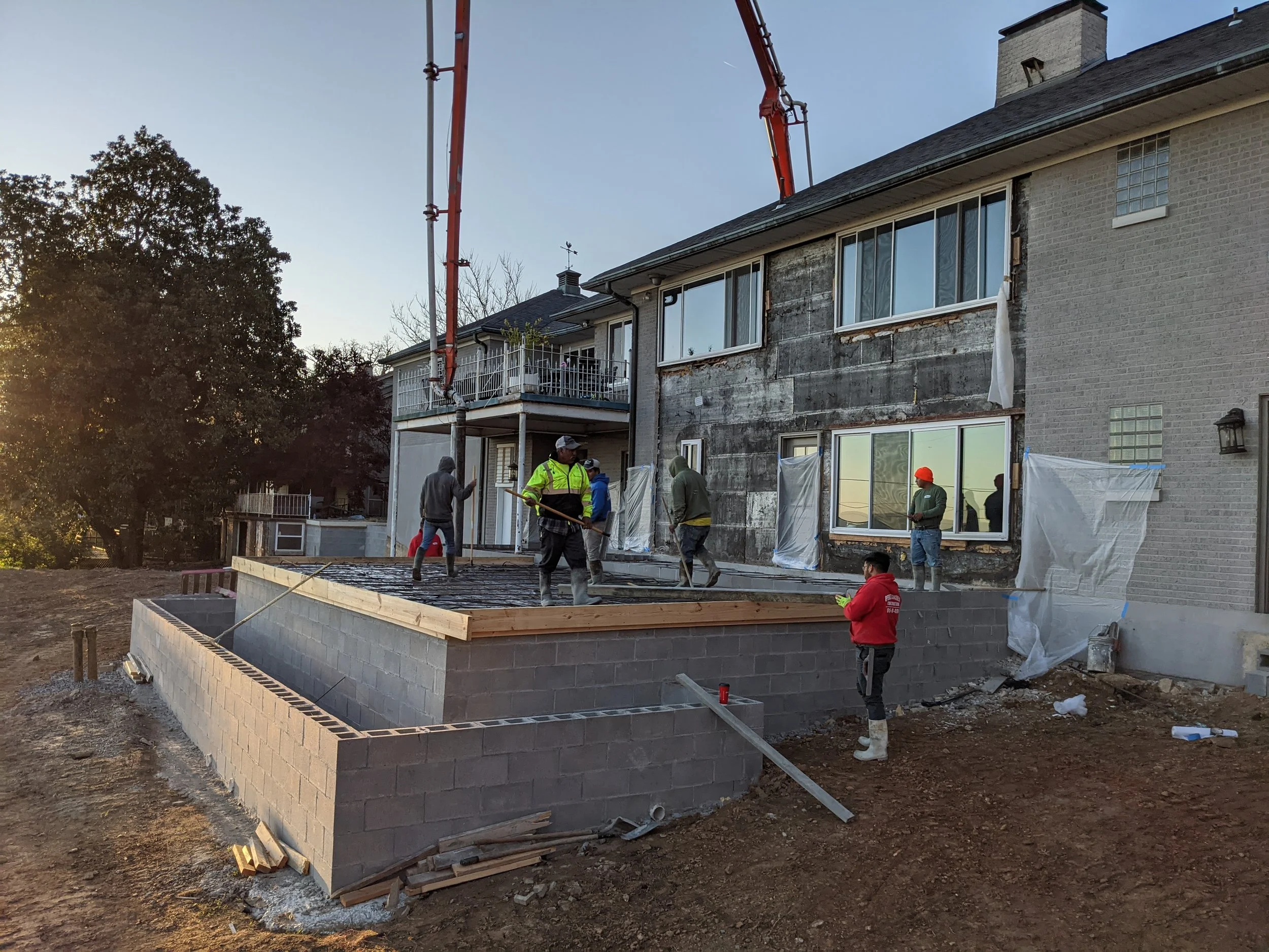 Construction workers building a house extension, with a brick foundation in progress, on a clear evening.