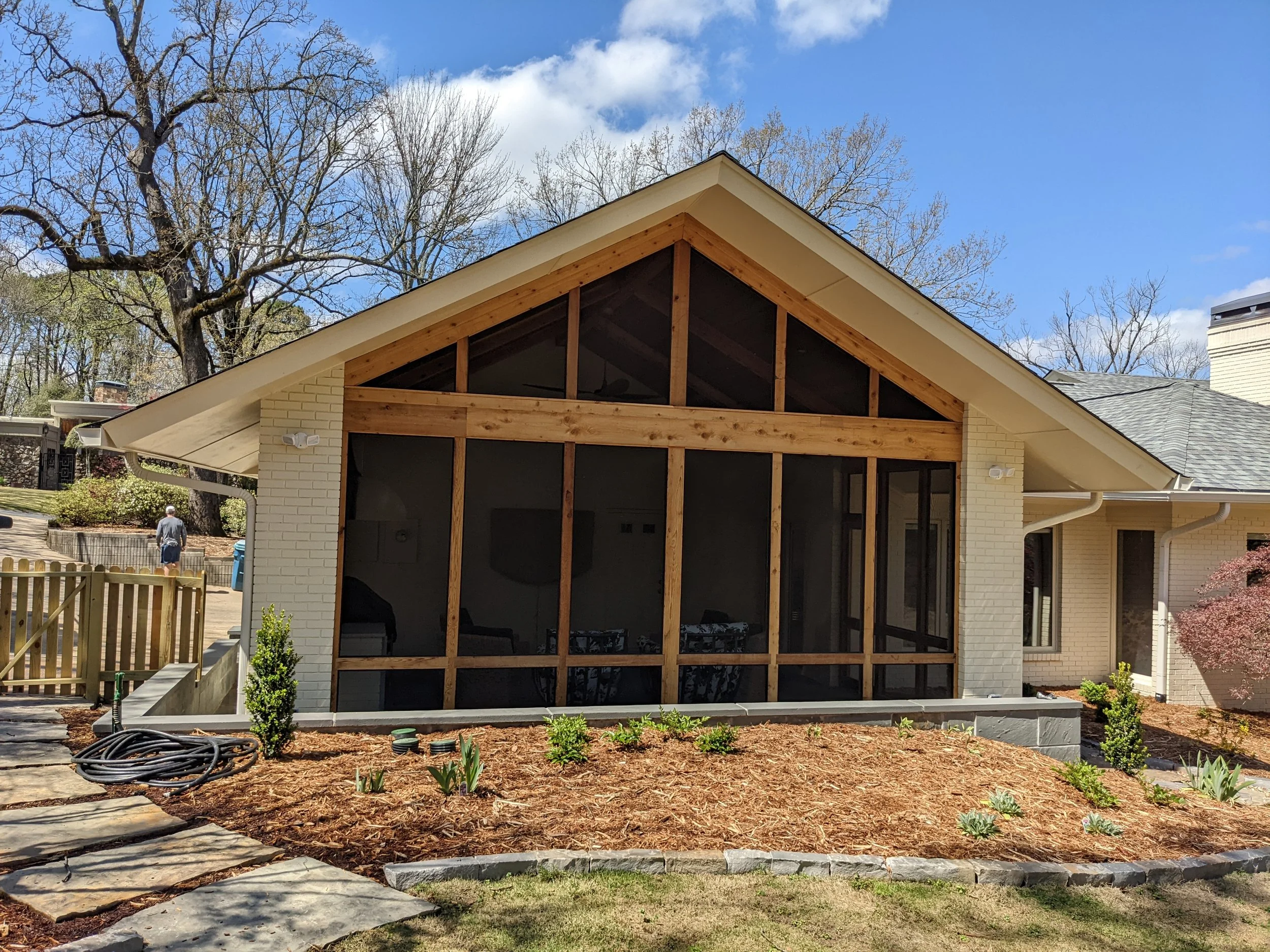 A house undergoing construction or renovation with a screened porch or sunroom addition being built. The house has a partially completed wooden frame for the new structure, with an angled roof and large windows. The yard has some small plants, mulch,