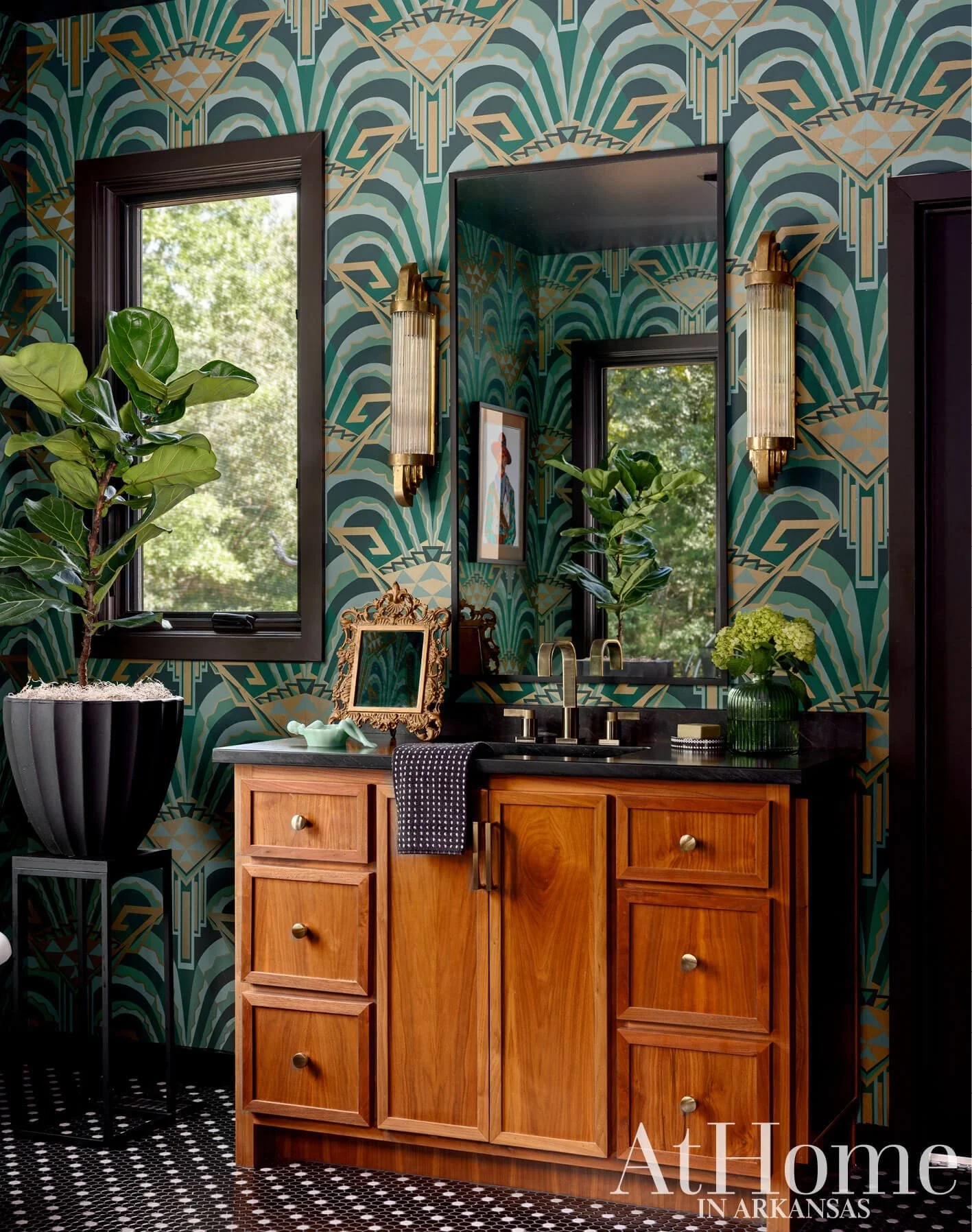 A bathroom with bold art deco patterned wallpaper in dark green, gold, and cream. There is a large window showing green trees outside. The mirror above the sink reflects the patterned wall, and is flanked by two vintage-style gold sconces. The black countertop has a faucet, a small framed picture, and some green plants in vases. The wooden vanity has six drawers with round metal knobs. The black and white mosaic tile floor completes the design.