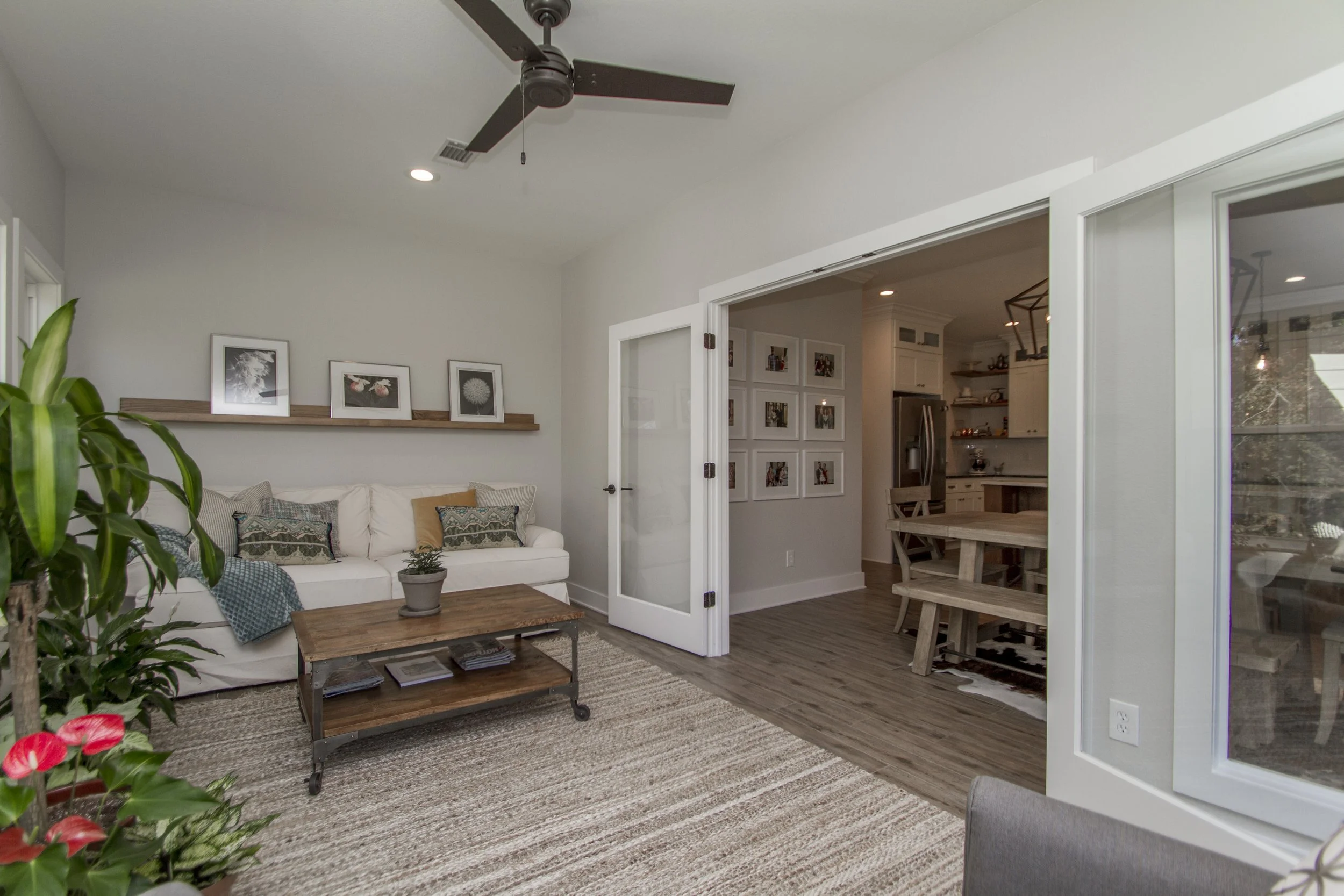 A living room with a white sofa, decorative cushions, a wooden coffee table, a framed picture shelf, a potted plant, and an open doorway leading to a dining area with a wooden table and chairs. Ceiling fan and recessed lighting visible.