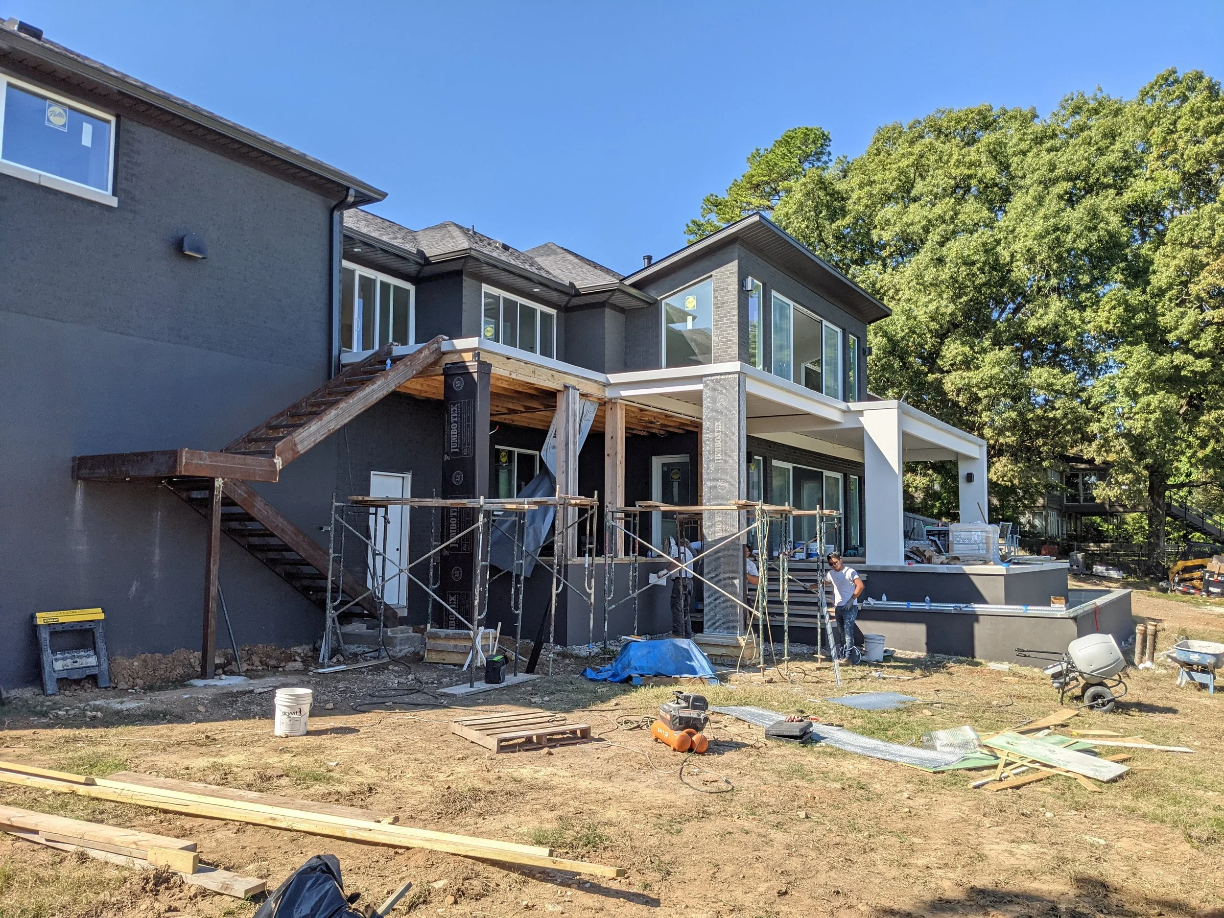 Construction site of a modern house, with workers, building materials, and scaffolding, under clear blue sky with trees in background.