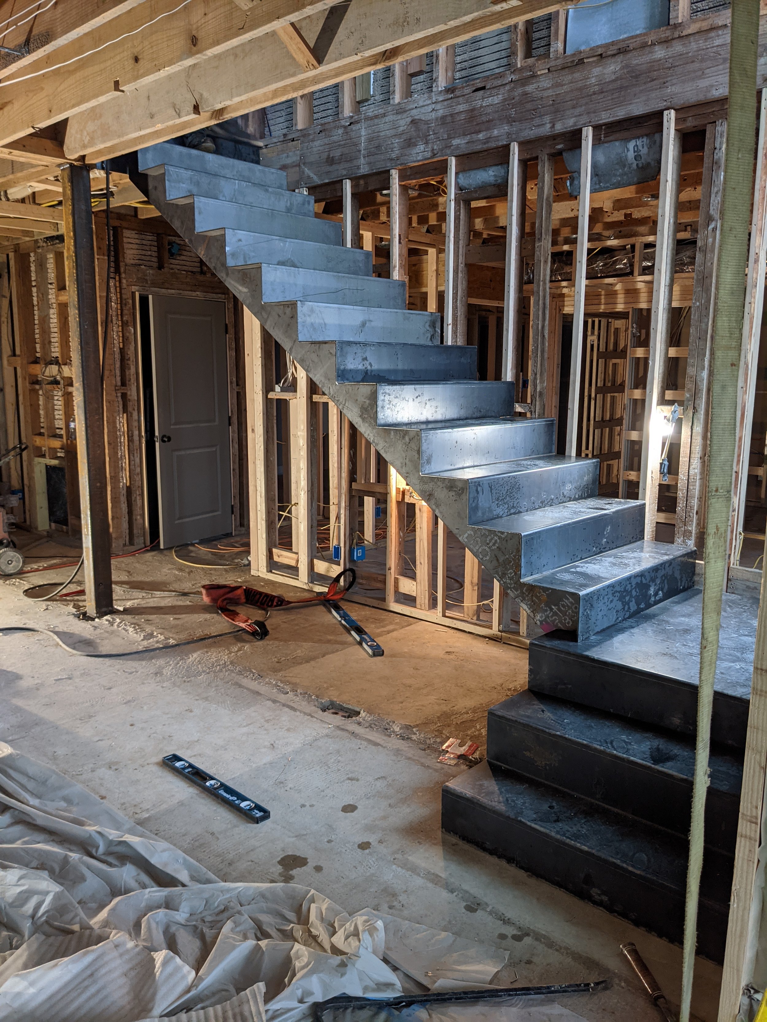 The image shows an unfinished interior of a building under construction with exposed wooden framing, a concrete staircase, construction tools, and materials scattered on the floor.