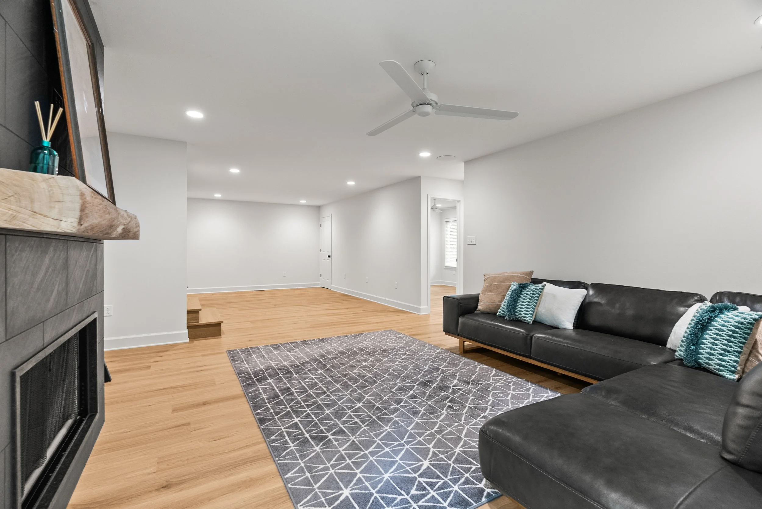 A living room with a black leather sofa decorated with blue and beige throw pillows, a gray patterned rug, white walls, hardwood flooring, a ceiling fan, and a window with blinds.
