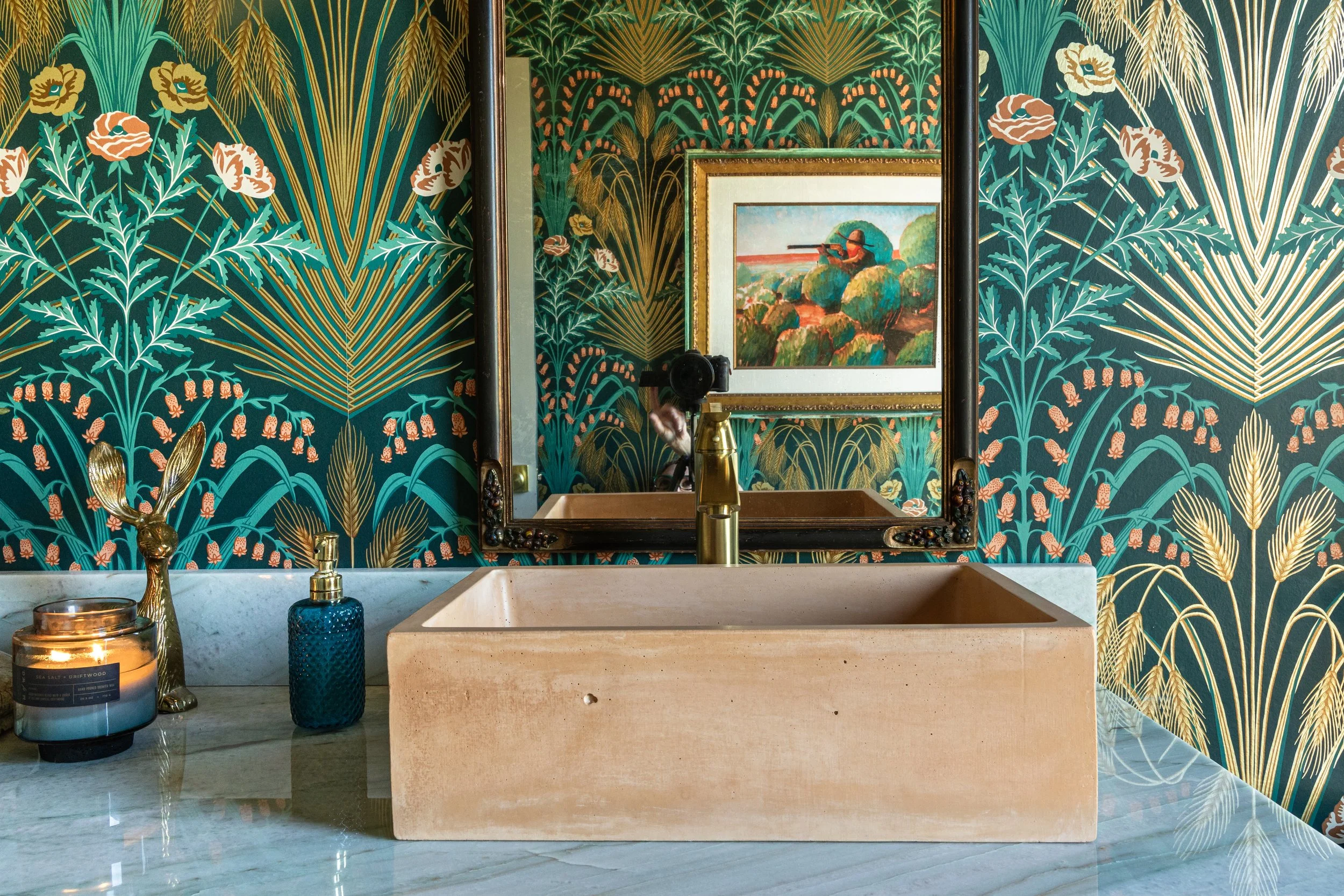 A bathroom sink area with a rectangular beige stone vessel sink on a marble countertop. Behind the sink is a large mirror framed in dark wood, reflecting a colorful landscape painting. The wallpaper has a lush, tropical pattern in dark green, gold, p